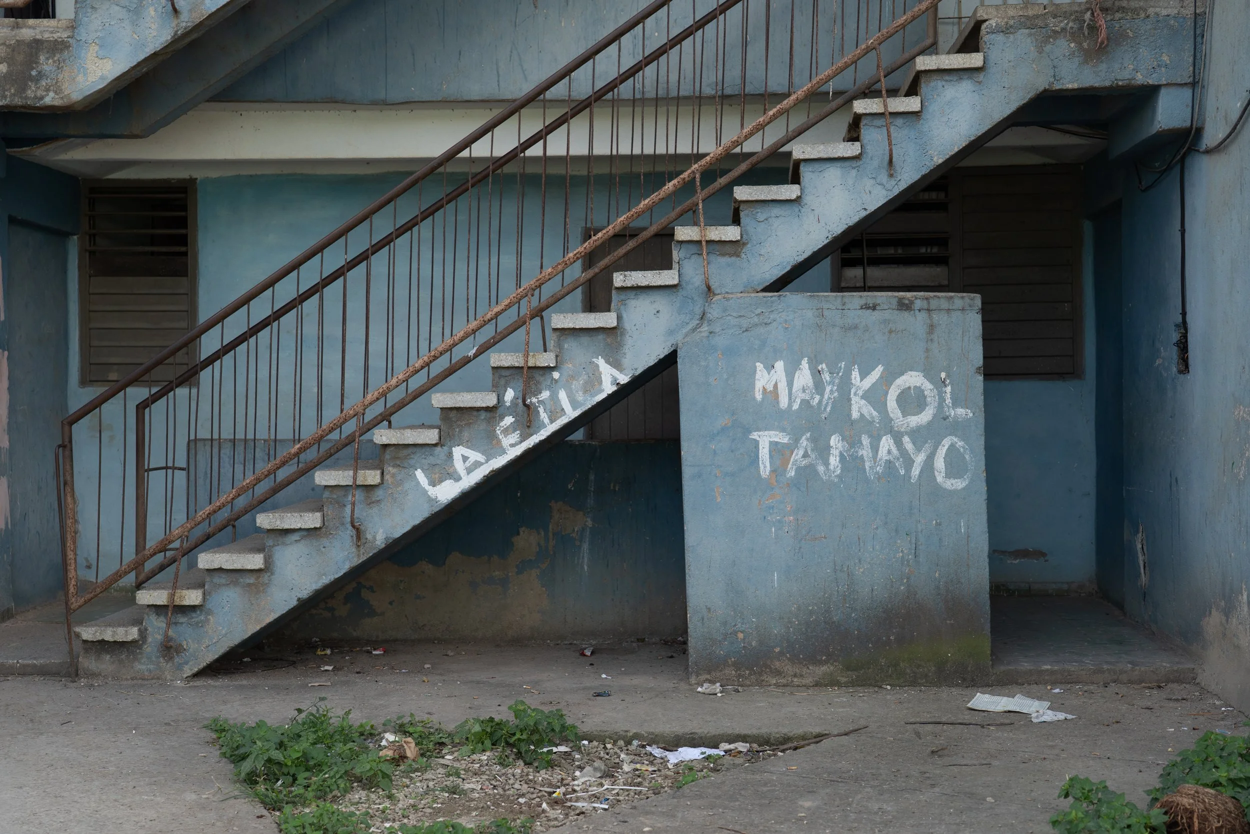 An outdoor concrete staircase with rusted metal railings leads to the second floor of a worn, blue building. Graffiti in white paint is visible on the wall beneath the stairs.