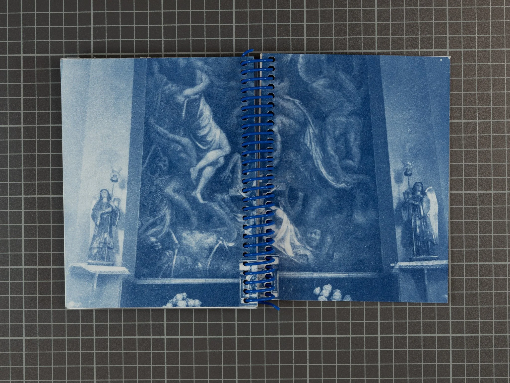 A blue-toned print of a religious painting inside a spiral-bound book, with two statues of saints on plinths on either side of the painting. The painting depicts a biblical scene with various figures, some kneeling and others standing, against a dark