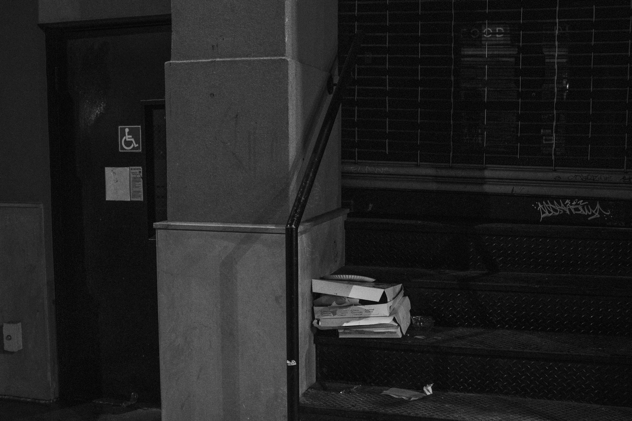 A set of black metal stairs with a few stacked pizza boxes and a plastic bottle on the landing. To the left, a concrete pillar and a small wall with a handicap accessible sign. The background shows a metal gate with graffiti.