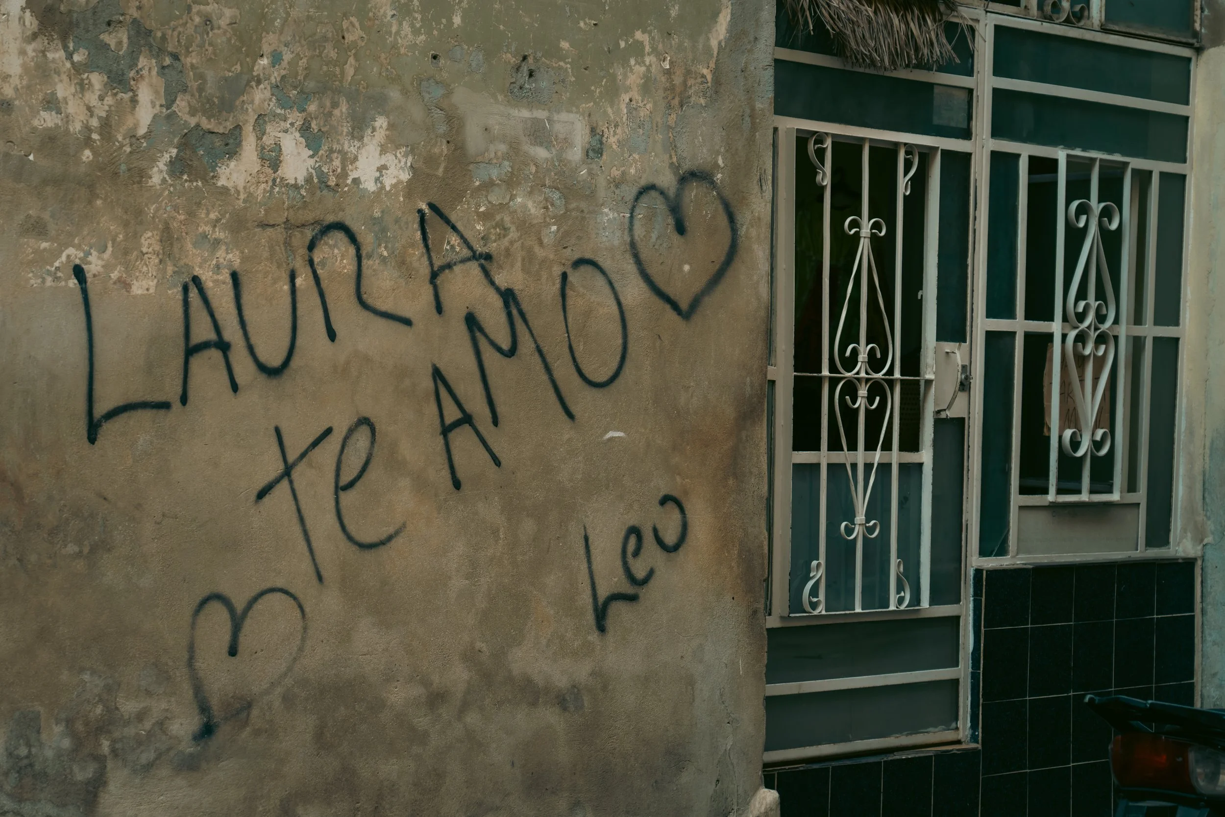 A wall with graffiti that reads 'LAURA te AMO' with a heart symbol, next to a window with decorative white metal bars.