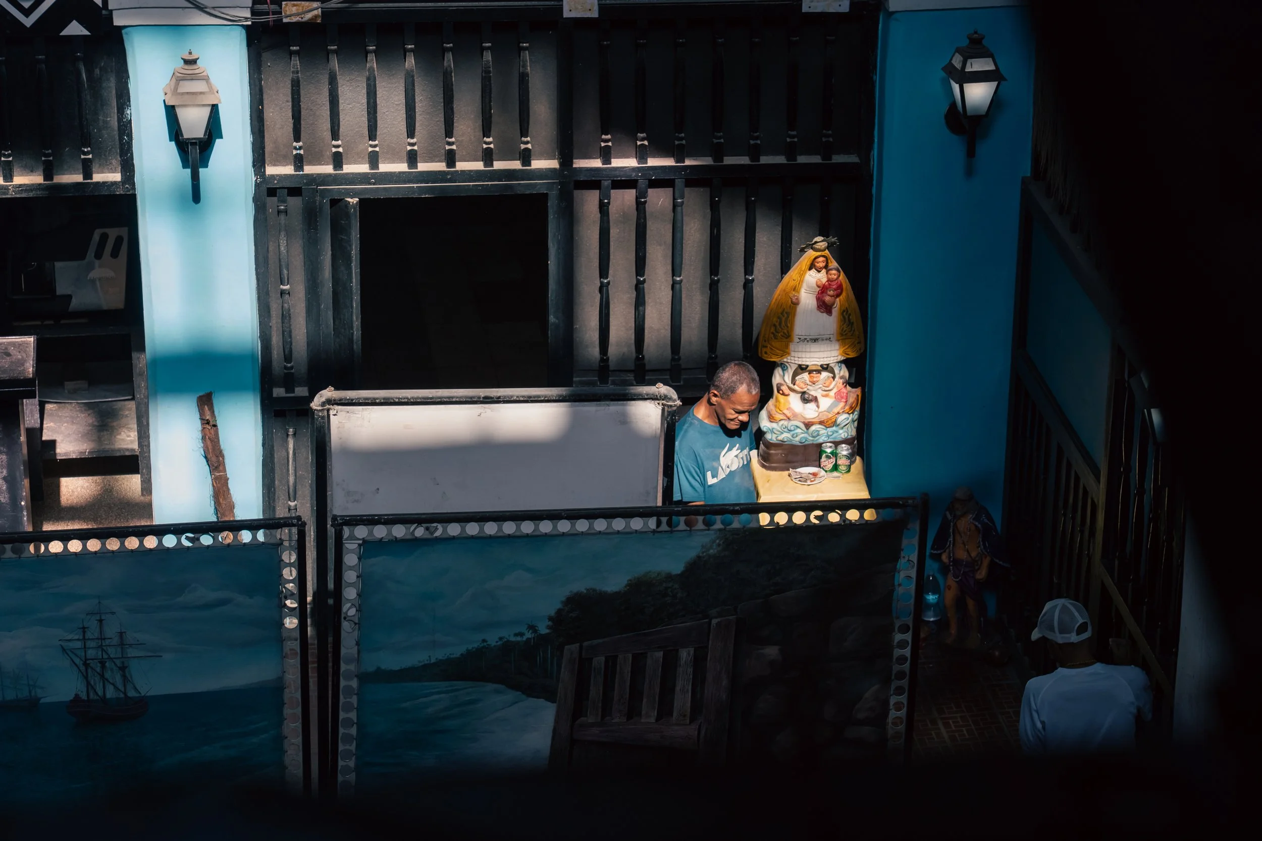 Man standing near religious statue and table with drink cans, in a small outdoor space with painted fences and lamps.