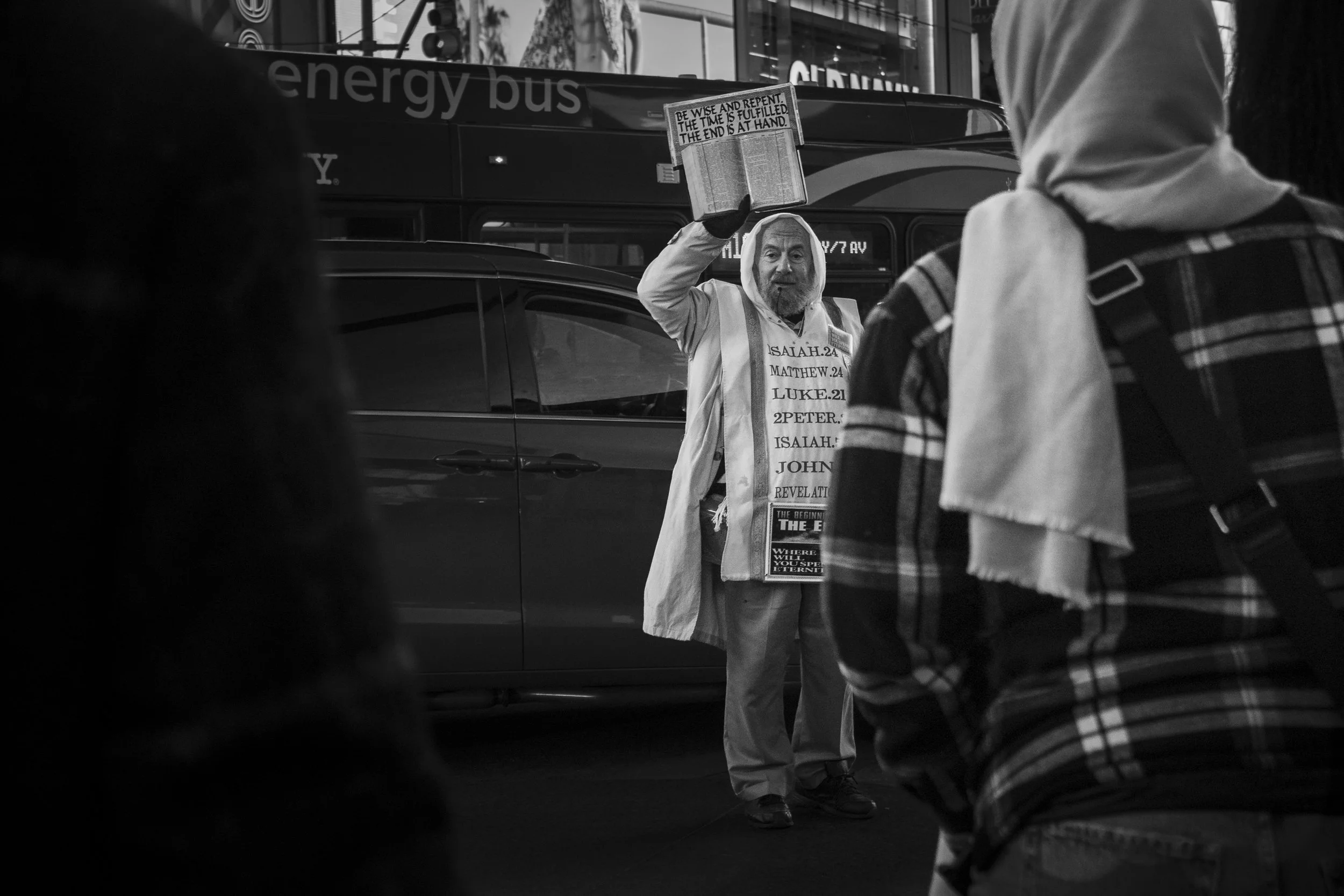 A man dressed in religious robes holding a sign with biblical references and reading from a book, standing near a car at a busy intersection, with two people in the foreground and a bus in the background.