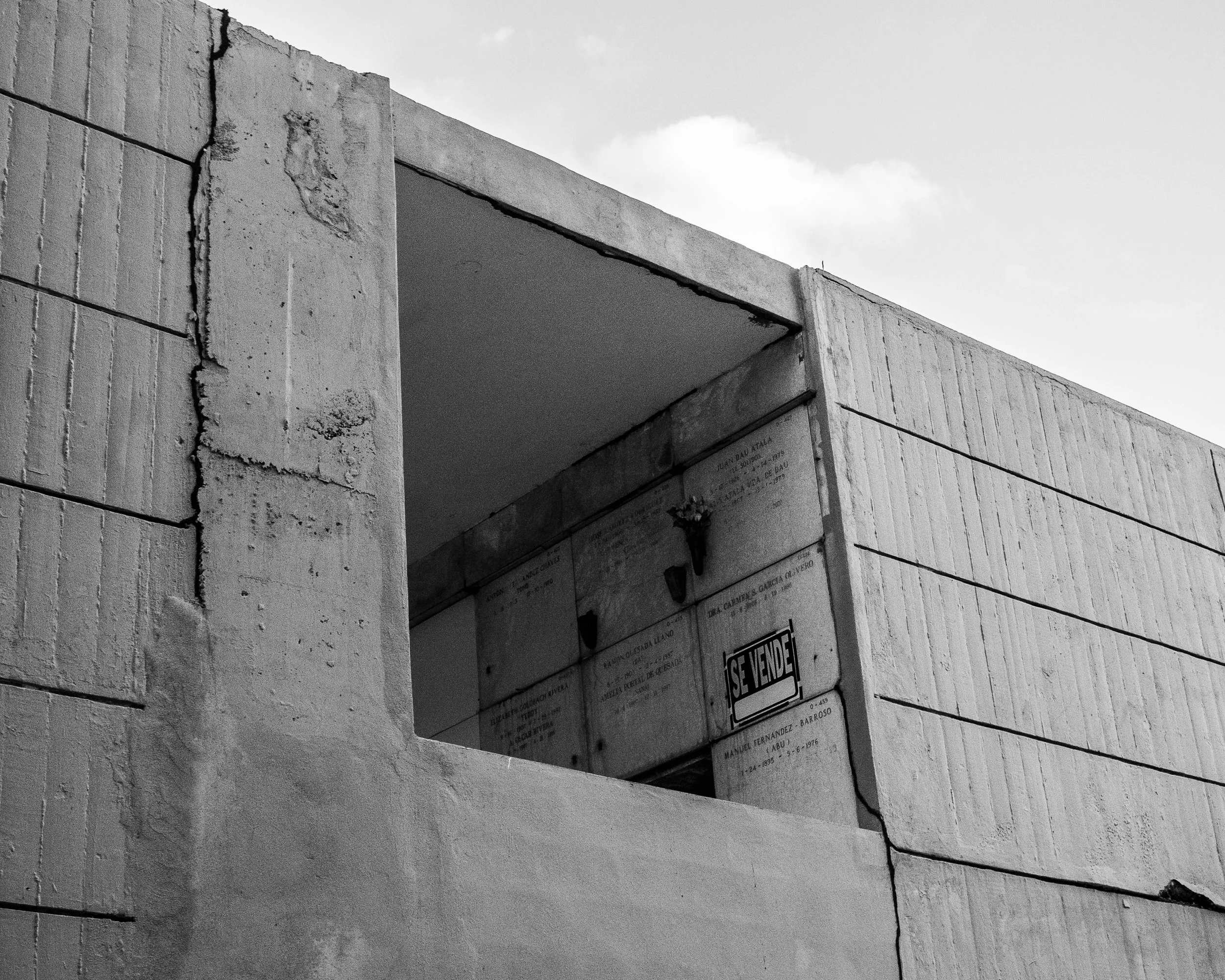 Black and white photograph of an above ground tomb concrete building with a large square opening and a sign that reads 'Se Vende' (For Sale) on the wall.