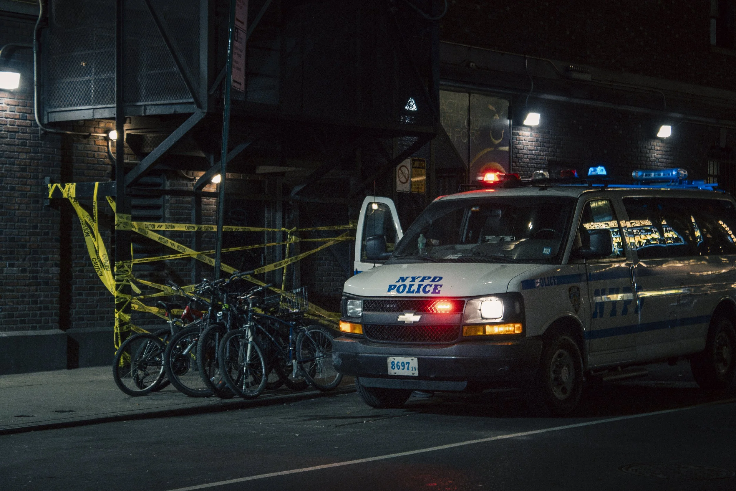 A NYPD police van with flashing red and blue lights parked on the street at night. Bicycles are chained to a bike rack, and yellow caution tape is wrapped around a nearby metal staircase, indicating a police investigation or scene.