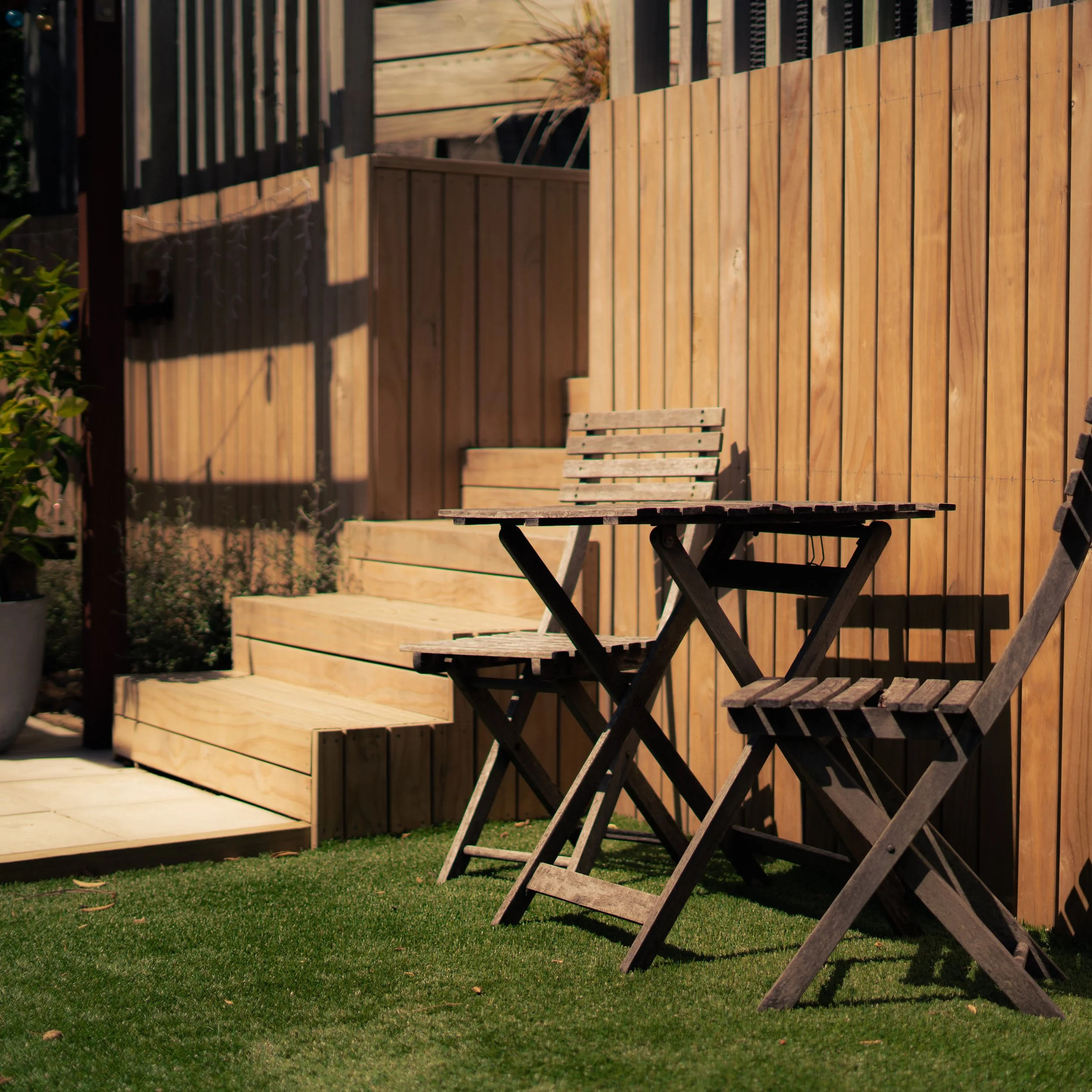 A small outdoor patio area with a wooden table and two matching chairs on a grassy lawn, enclosed by a tall wooden fence, with stairs leading up to a raised platform in the background.