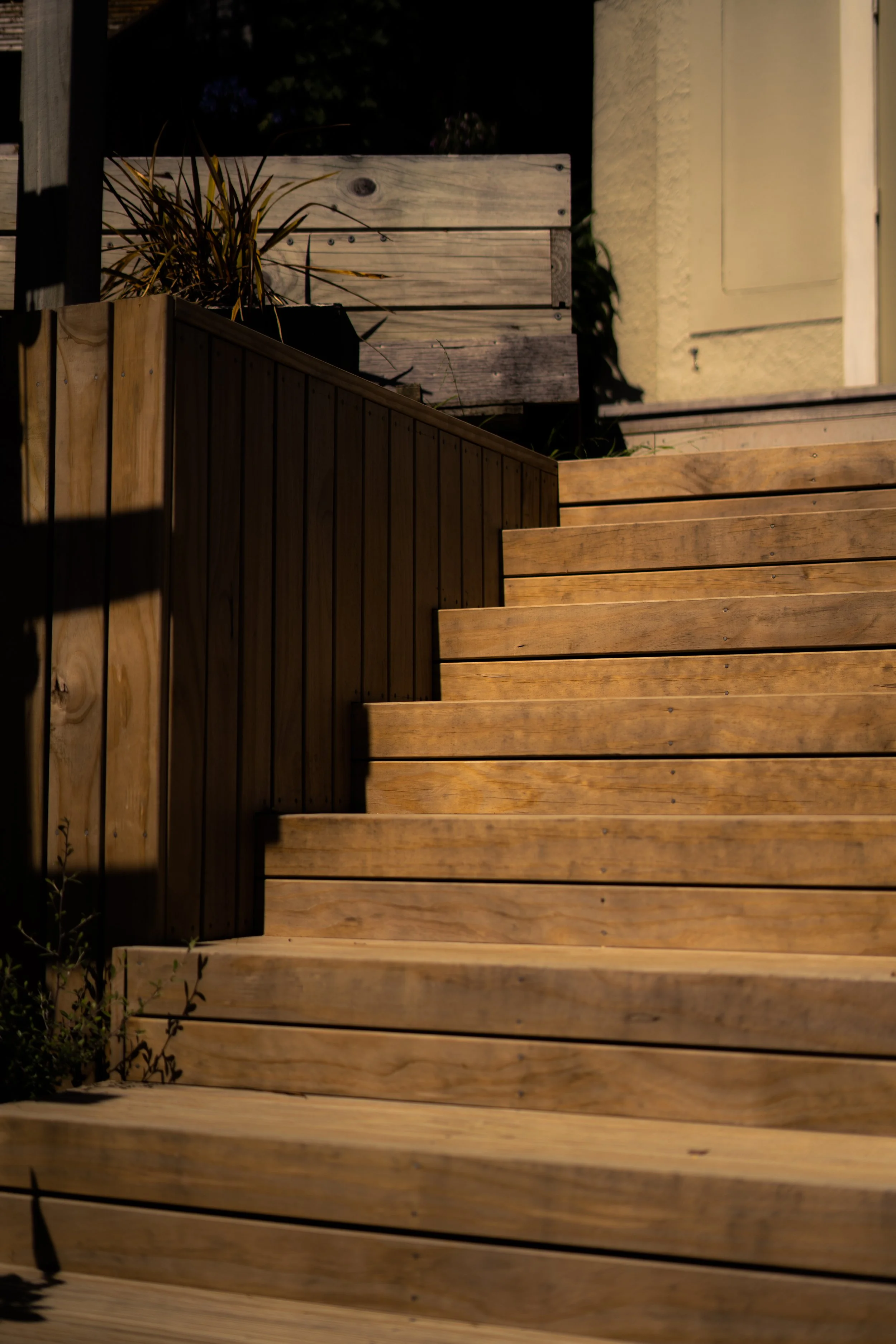 Wooden stairs leading up to a house entrance, with potted plants and shadows.