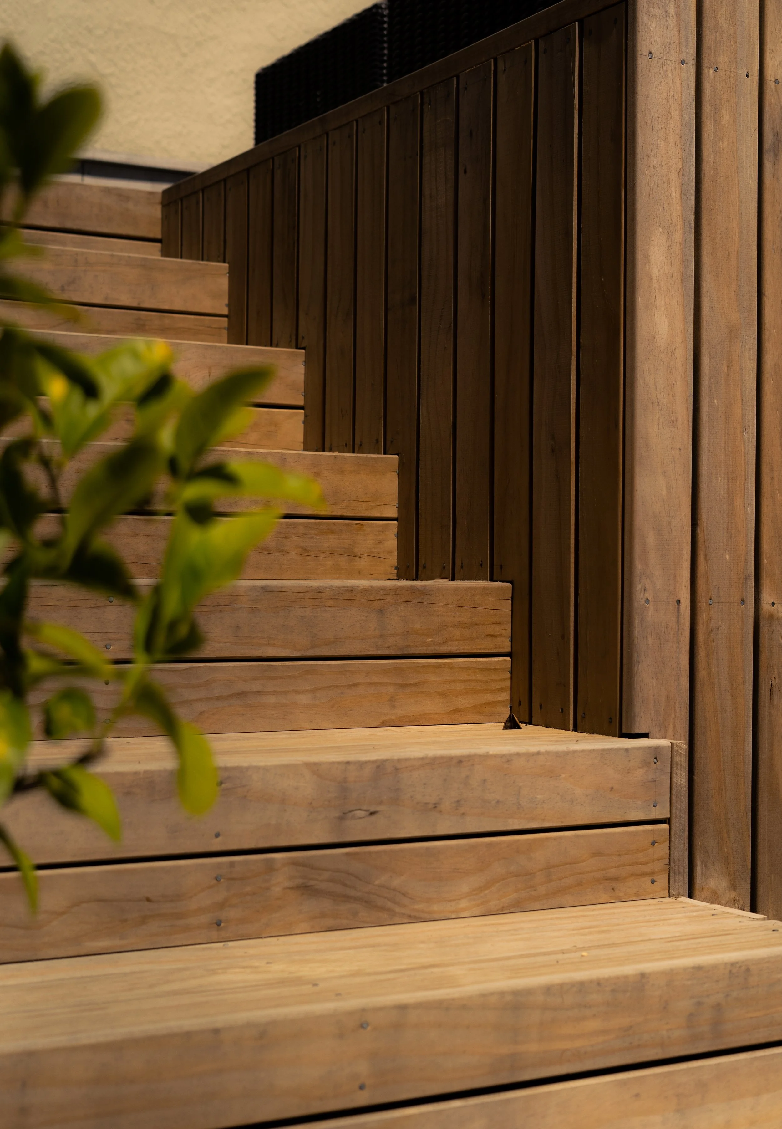 Wooden outdoor staircase with horizontal planks and a wooden privacy fence in the background, partially obscured by green leaves in the foreground.