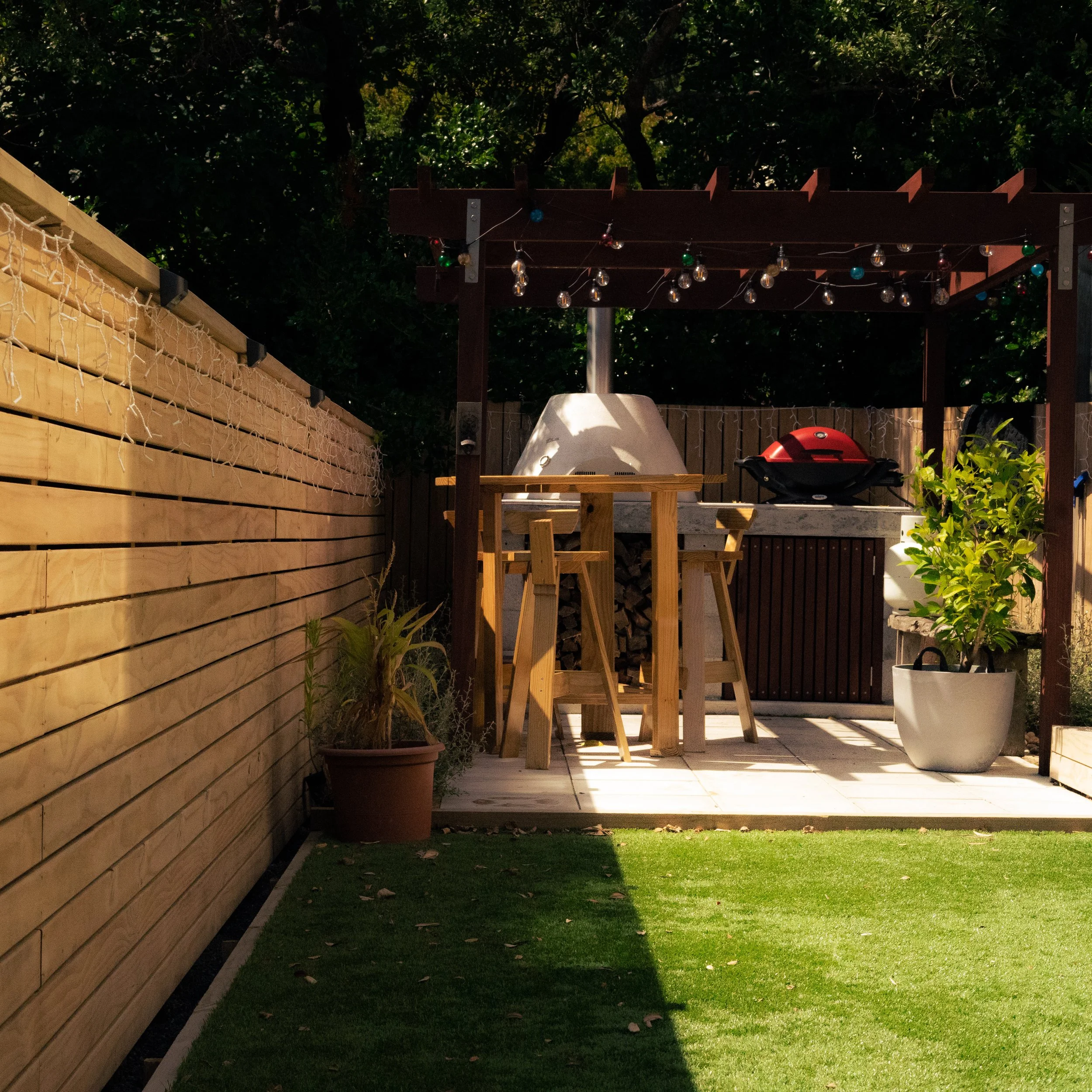 Backyard patio area with a grill station, wooden bar table and chairs under a string light covered pergola, surrounded by a wooden privacy fence, potted plants, and a grassy lawn.
