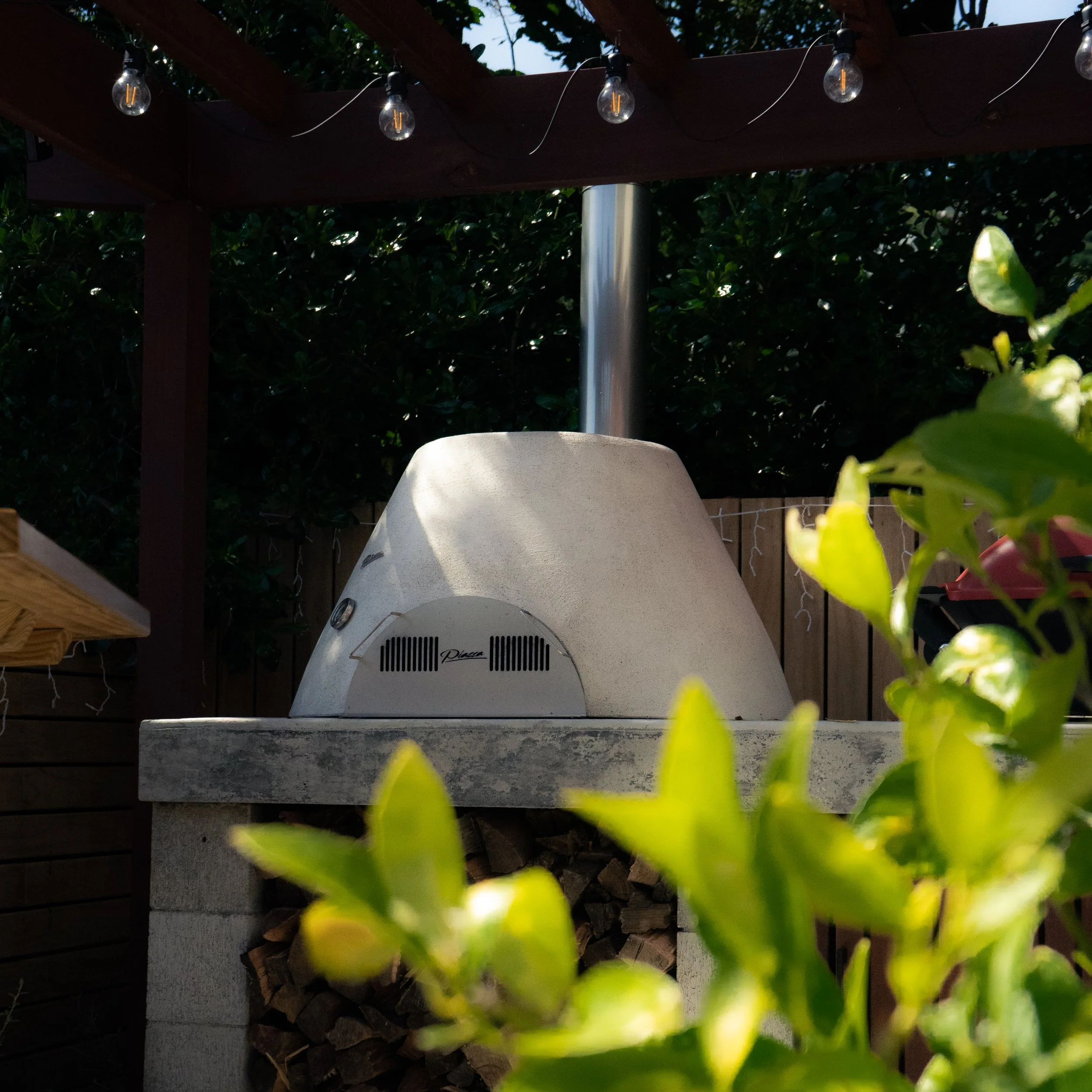 Outdoor pizza oven under string lights with green foliage in the background