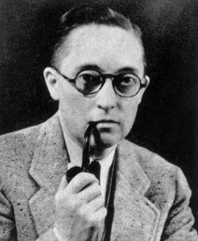 A young man wearing round glasses and a suit, holding a pipe in his hand.