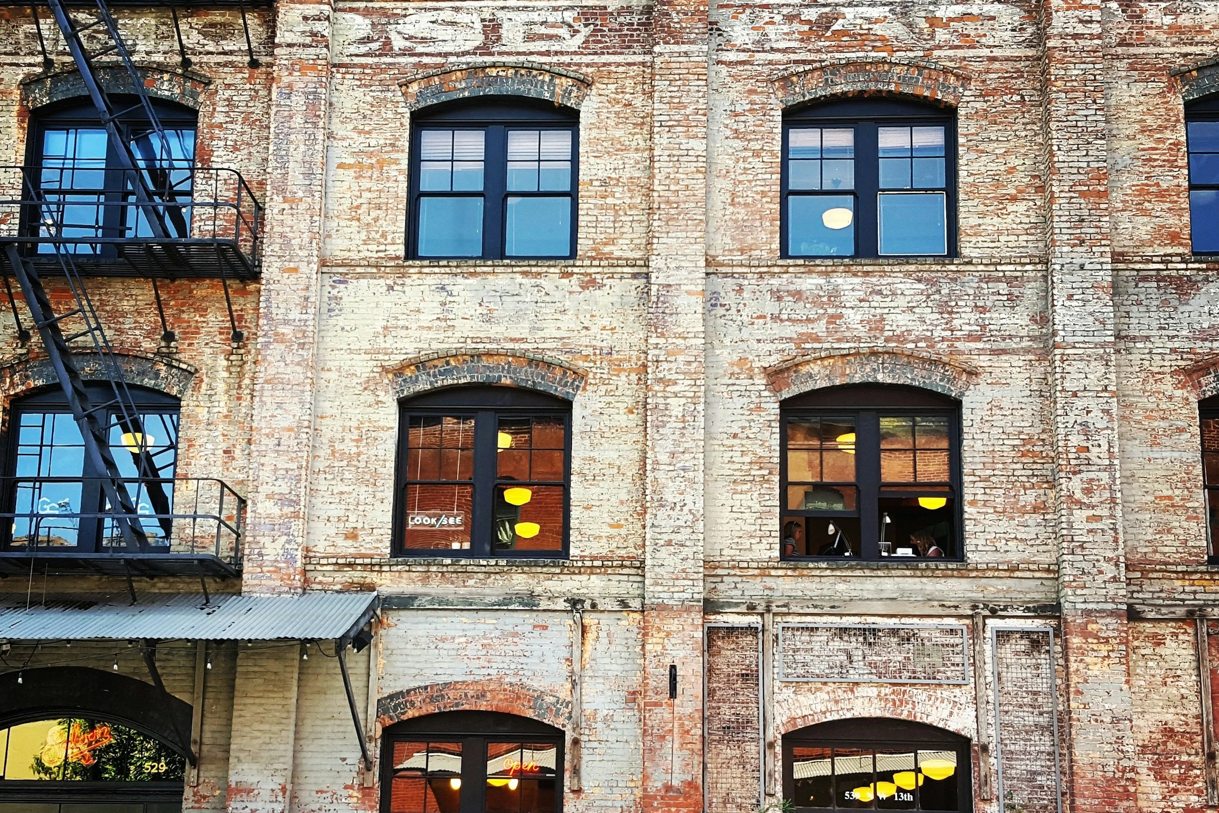 Brick building with multiple black-framed windows, some with yellow and white lights inside, and a black fire escape on the left side.