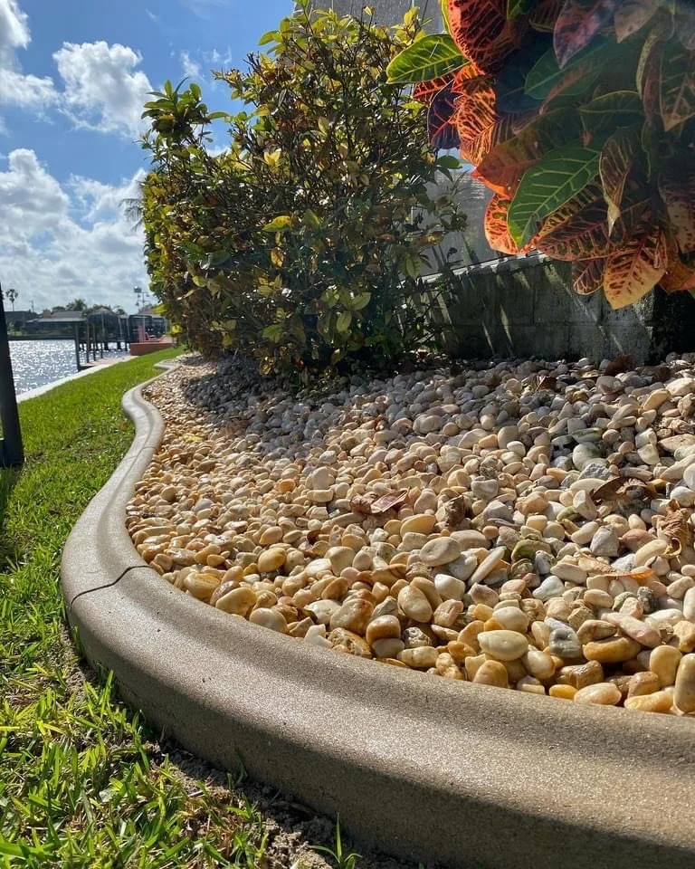 A landscaped garden bed edged with a concrete curbing, filled with small multicolored river rocks and green bushes, under a partly cloudy sky beside a waterway.