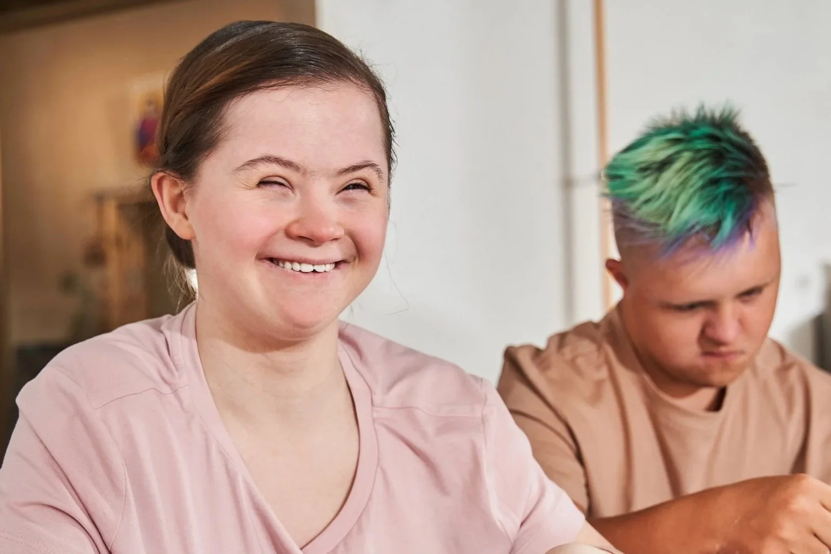 A young woman smiling happily with a person with rainbow-colored hair in the background.