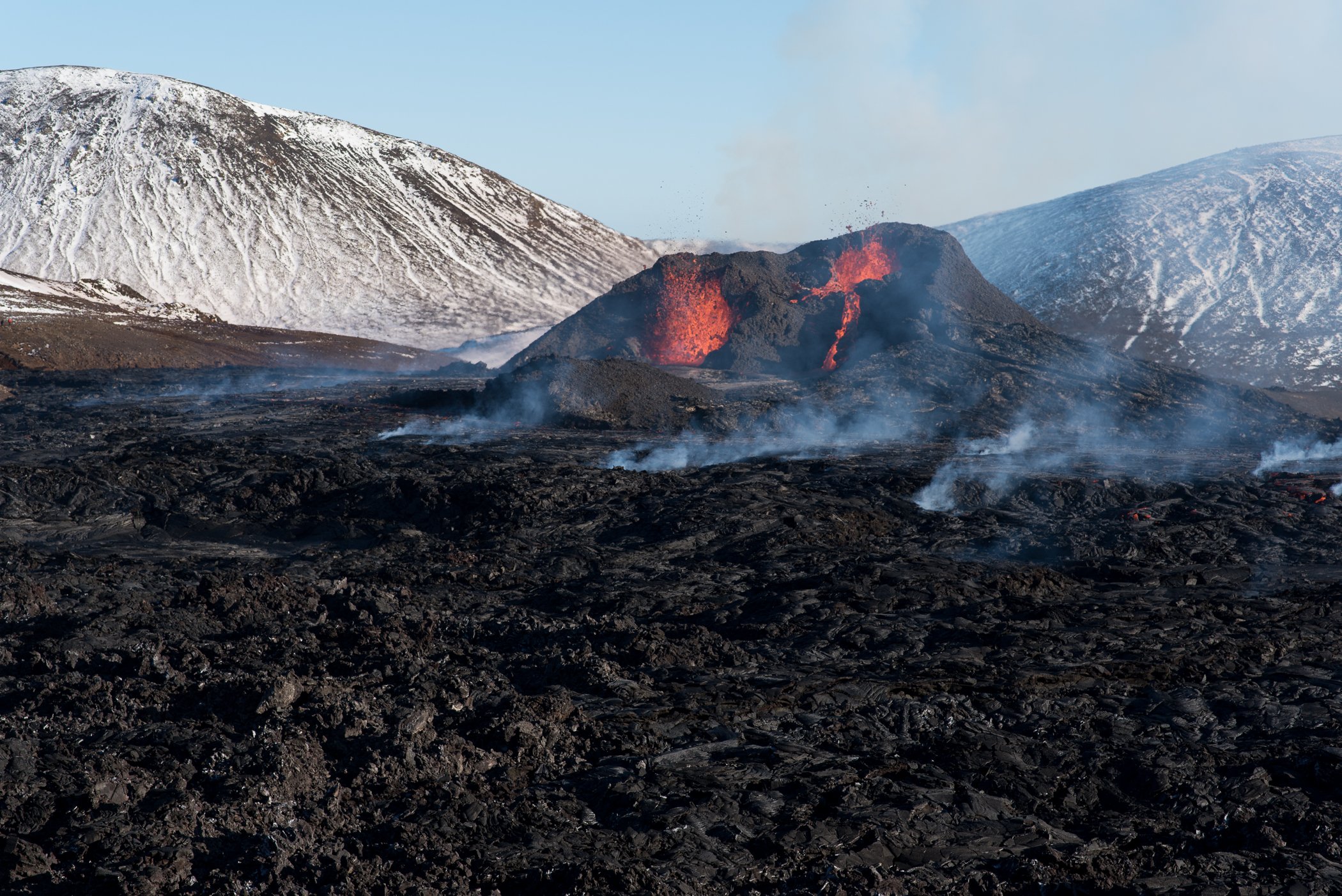 Volcanic eruption with red lava and smoke on a black rocky landscape, snowy mountains in the background.