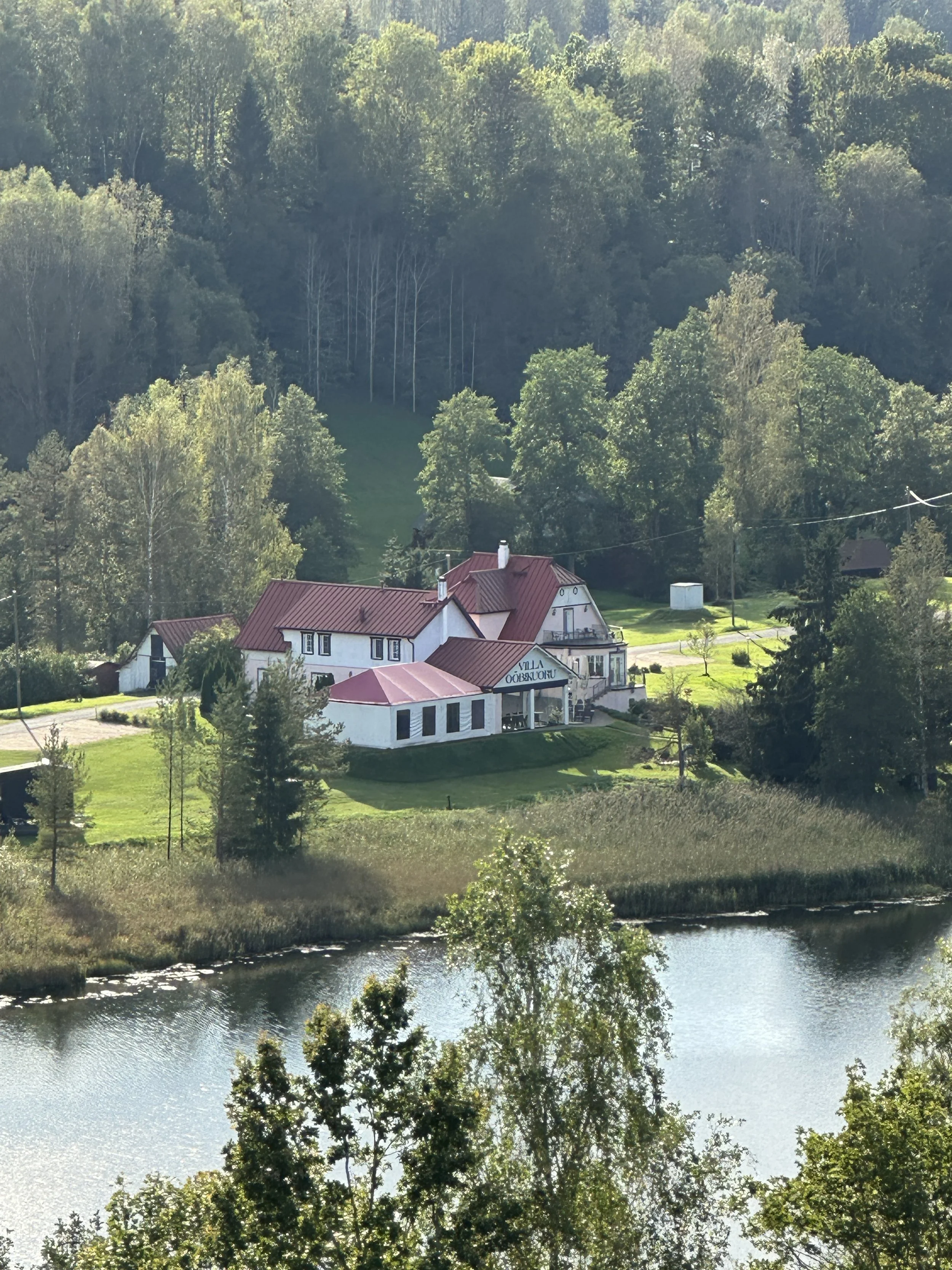 A large white house with a red roof situated on a grassy hillside next to a body of water, surrounded by trees and lush greenery.