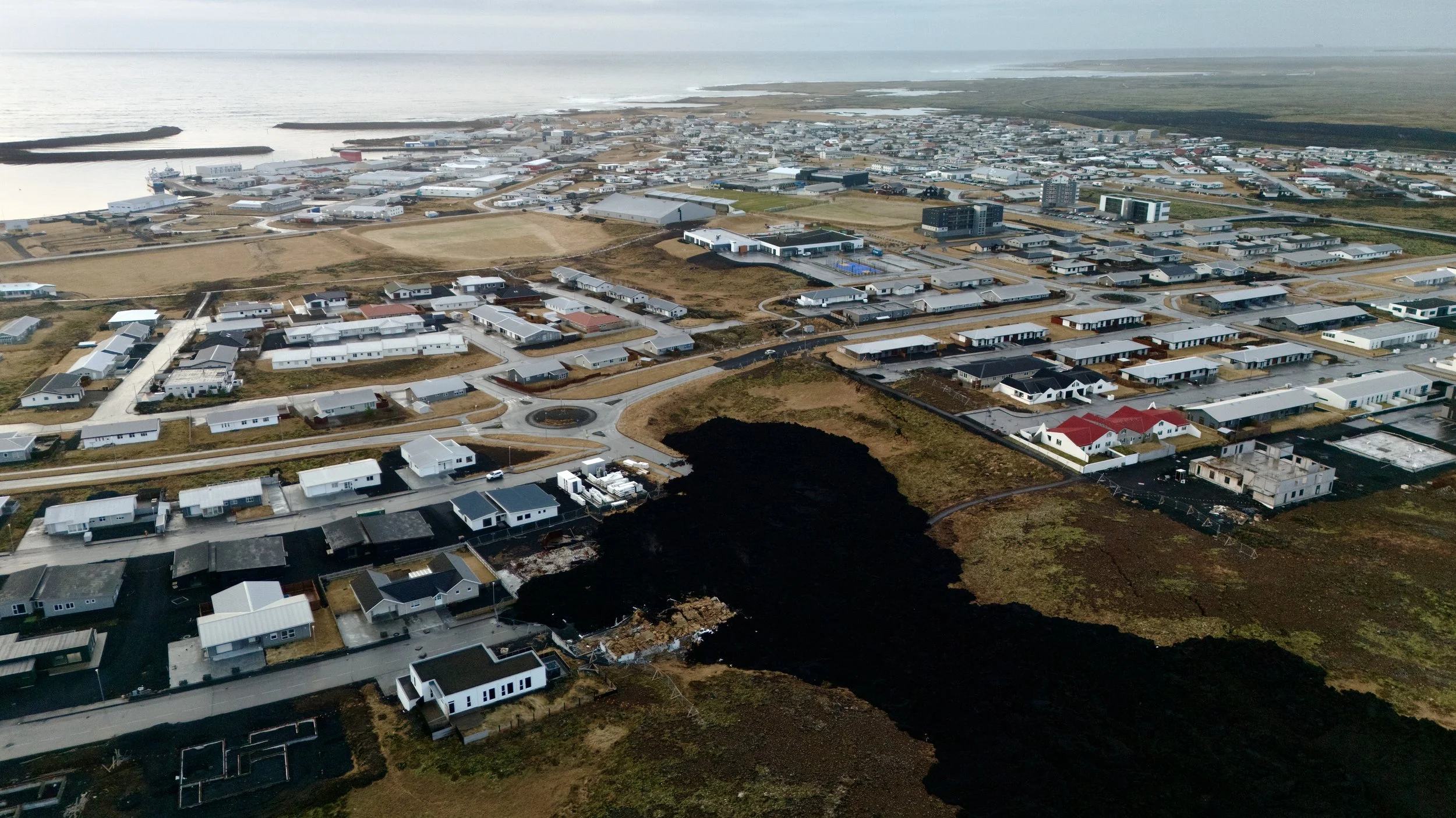 Aerial view of a residential area near the coast with houses, roads, and a black volcanic hill in the foreground.