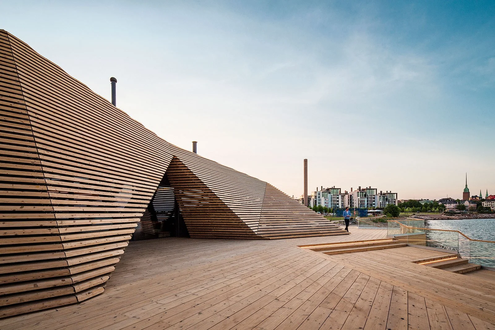 Modern wooden deck with geometric architectural structure overlooking a river with city buildings in the background