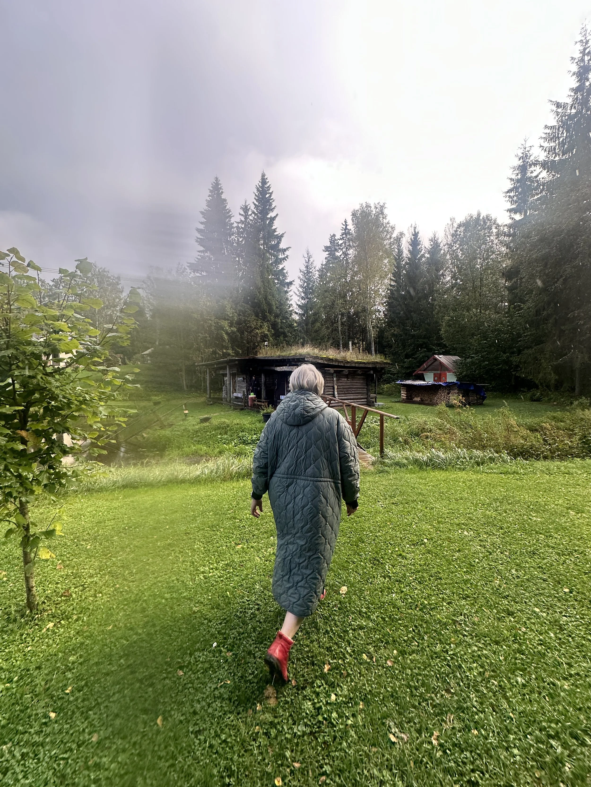 A person with gray hair, wearing a quilted coat and red shoes, walking towards a rustic wooden cabin in a green outdoor setting, surrounded by trees and grass.
