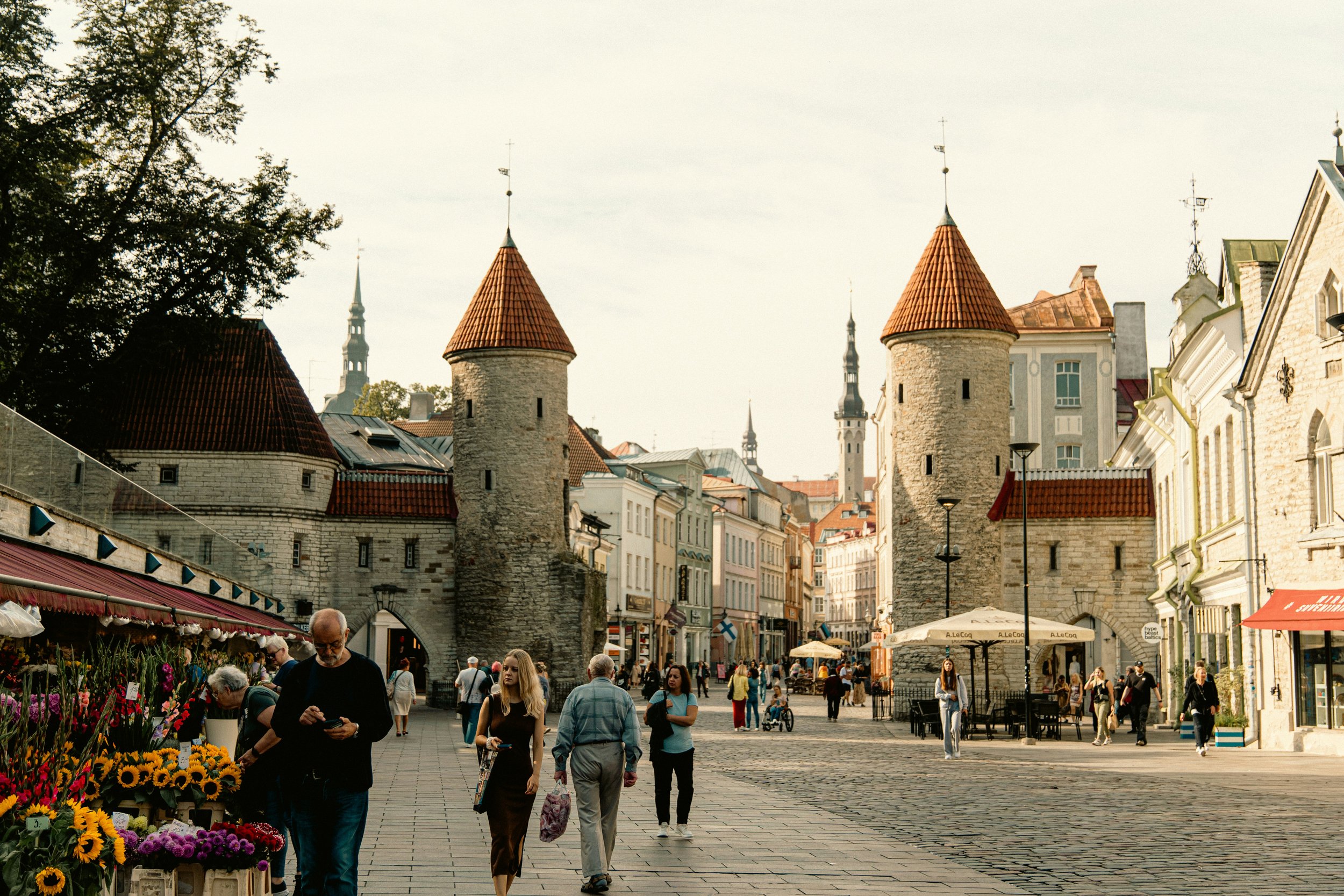 People walking in a historic city square with medieval stone towers, shops, and outdoor cafes.