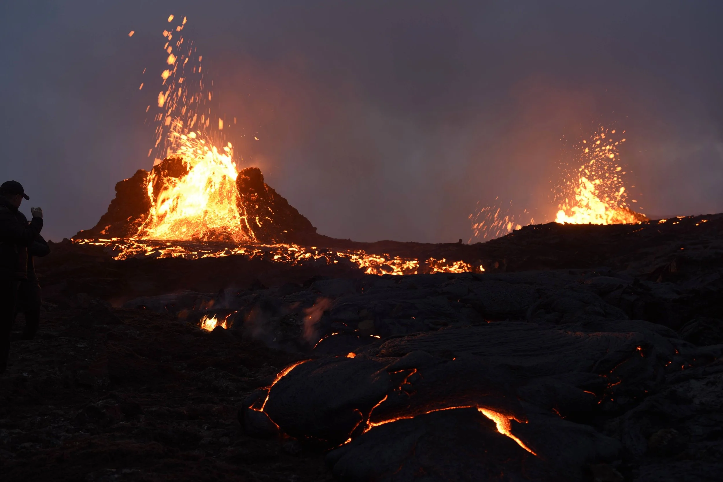 Eruption of two volcanoes with flowing lava, glowing rocks, and ash in the dark sky, with a person taking a photo on the left side.