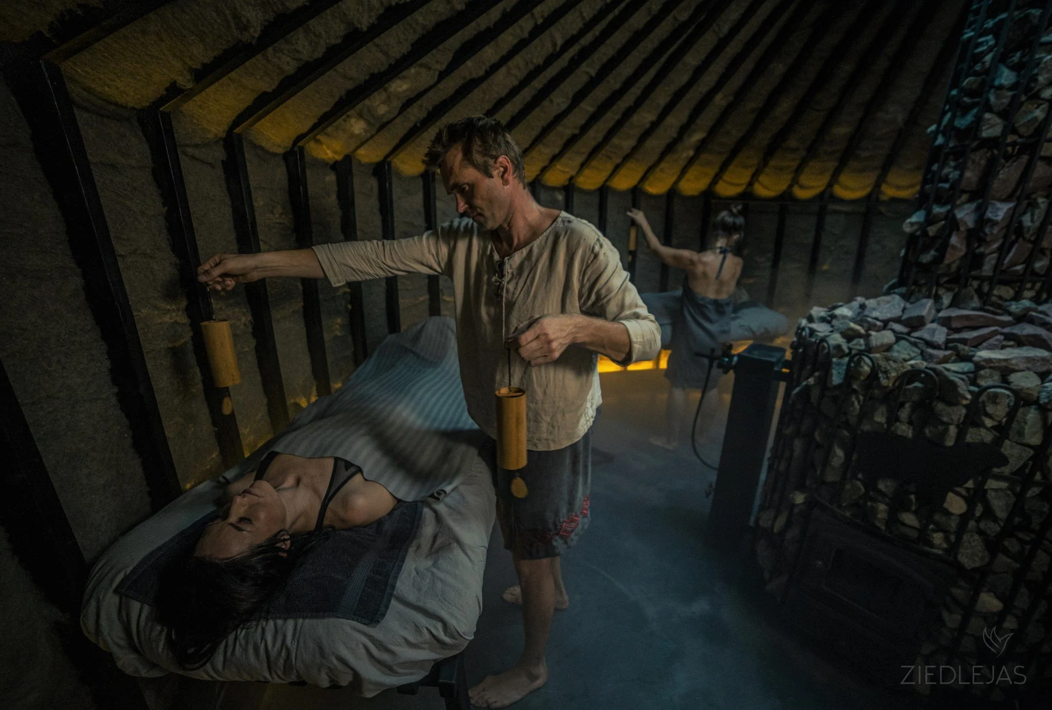 A man receiving a massage lying on a bed inside a rustic, dimly lit room with stone walls and a thatched ceiling, with a woman in the background attending to another person.