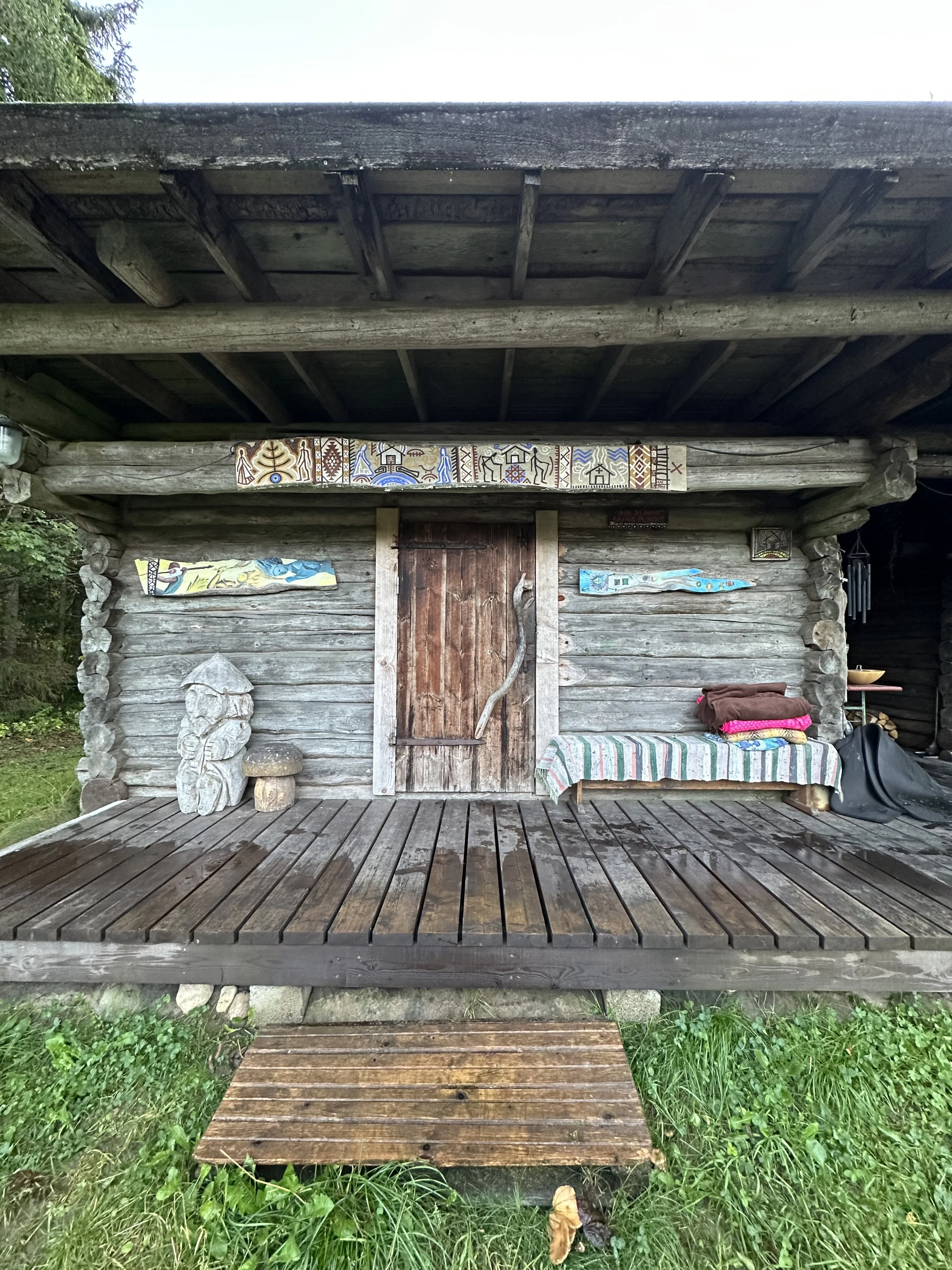 Wooden cabin with stone accents, decorated with colorful artwork and carvings, on a small wooden porch surrounded by grass.