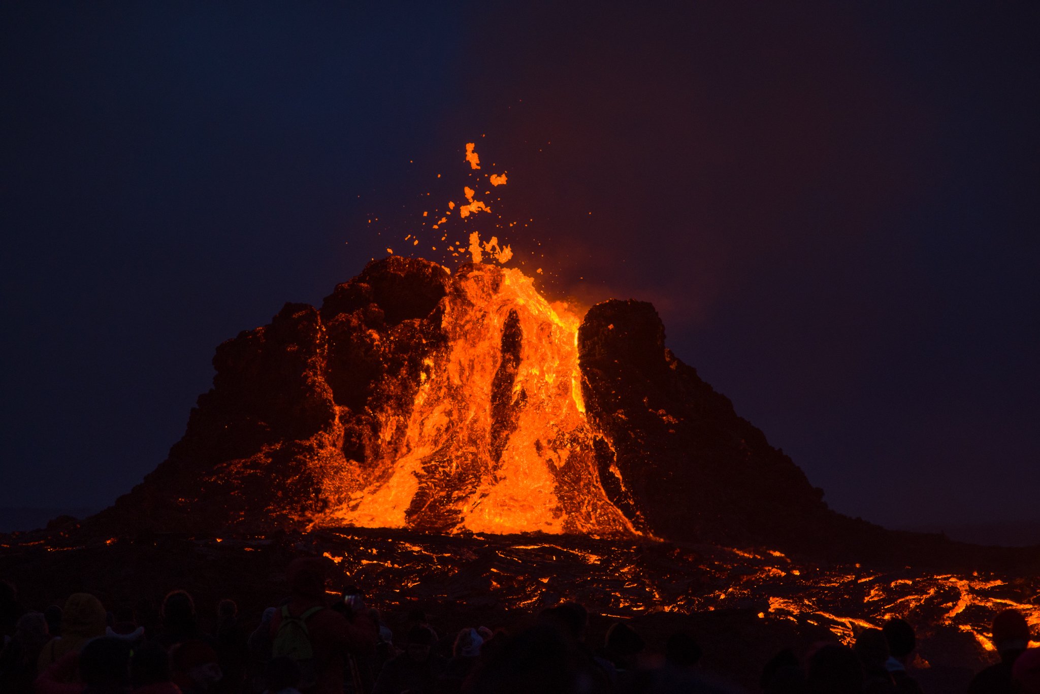 Erupting volcano spewing lava at night, with a crowd of people watching in the foreground.