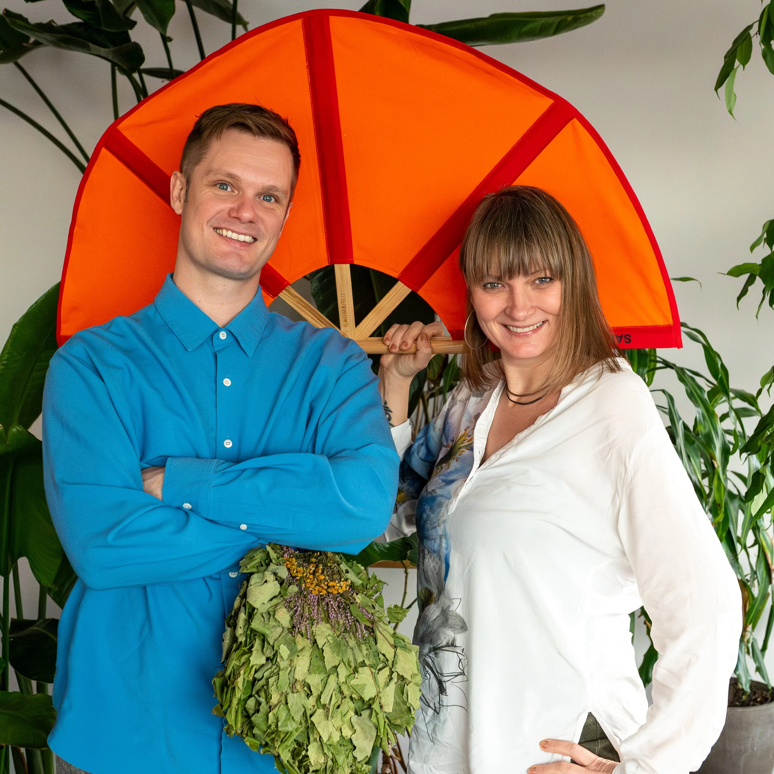 A man and woman standing indoors with green plants in the background. The man is wearing a blue shirt and is holding a plant with green leaves and purple flowers. The woman is wearing a white shirt and holding a large orange and red umbrella over their heads, smiling.