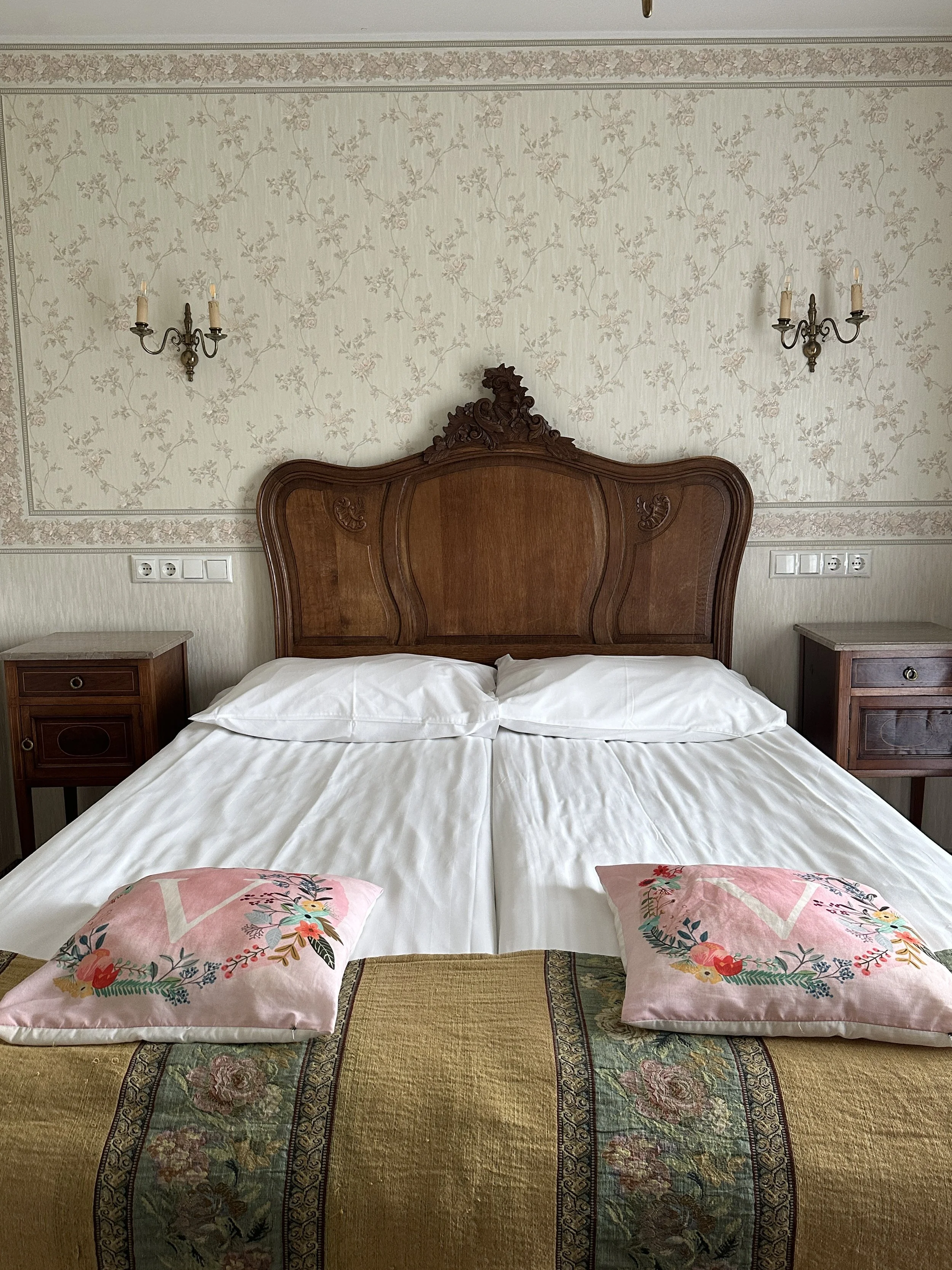 A bedroom with a large bed, ornate wooden headboard, two matching bedside tables, floral patterned lampshades, vintage wallpaper, and decorative wall sconces.