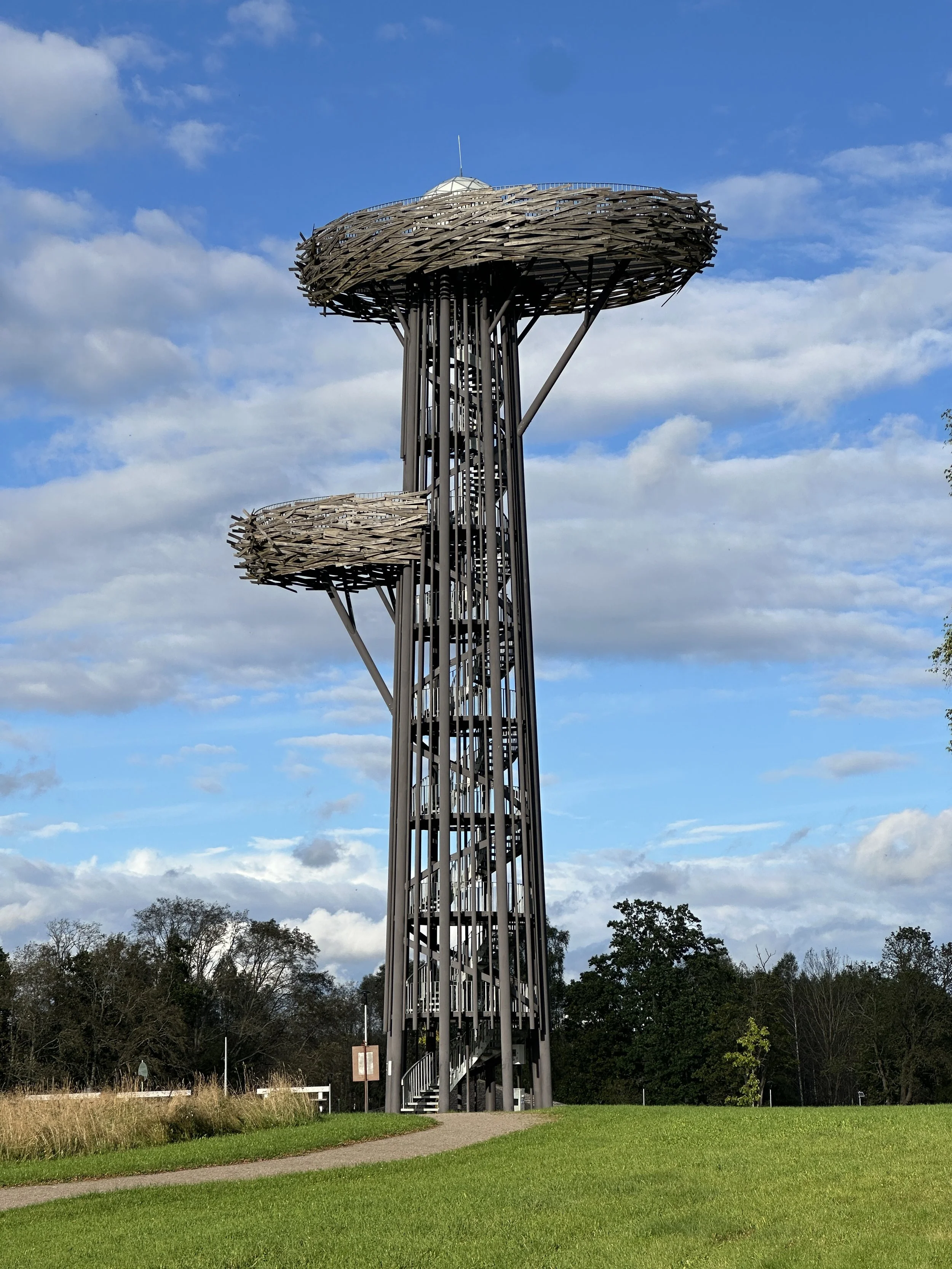 A tall fire lookout tower with a unique design, featuring two large, circular observation decks with wooden lattice roofs and a spiral staircase inside, set in a grassy area with trees in the background under a partly cloudy sky.