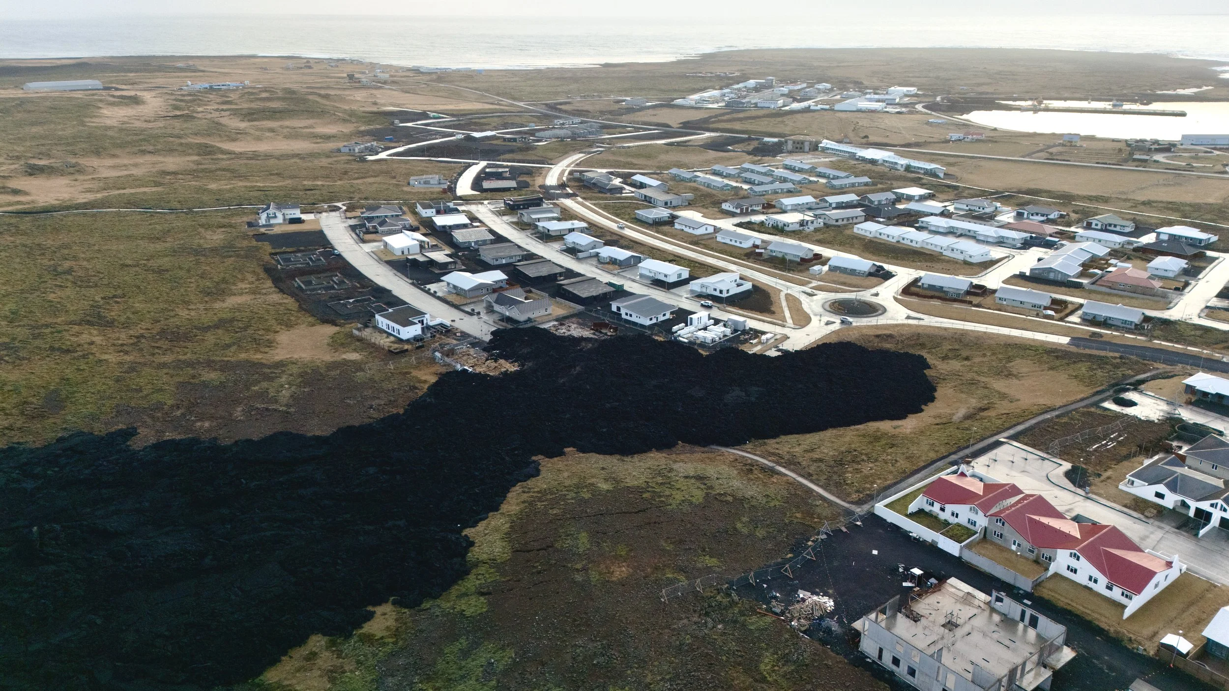 Aerial view of a residential neighborhood near a coastline, with multiple houses, roads, and some volcanic lava flow extending through part of the area.