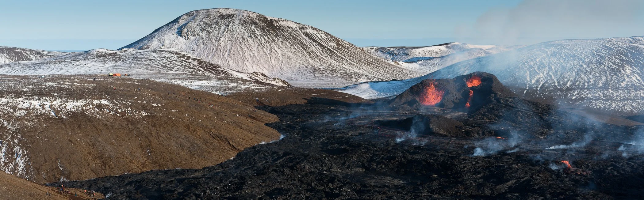 Active volcano with flowing lava, surrounded by snow-capped mountains and land, with visible smoke and ash emissions.