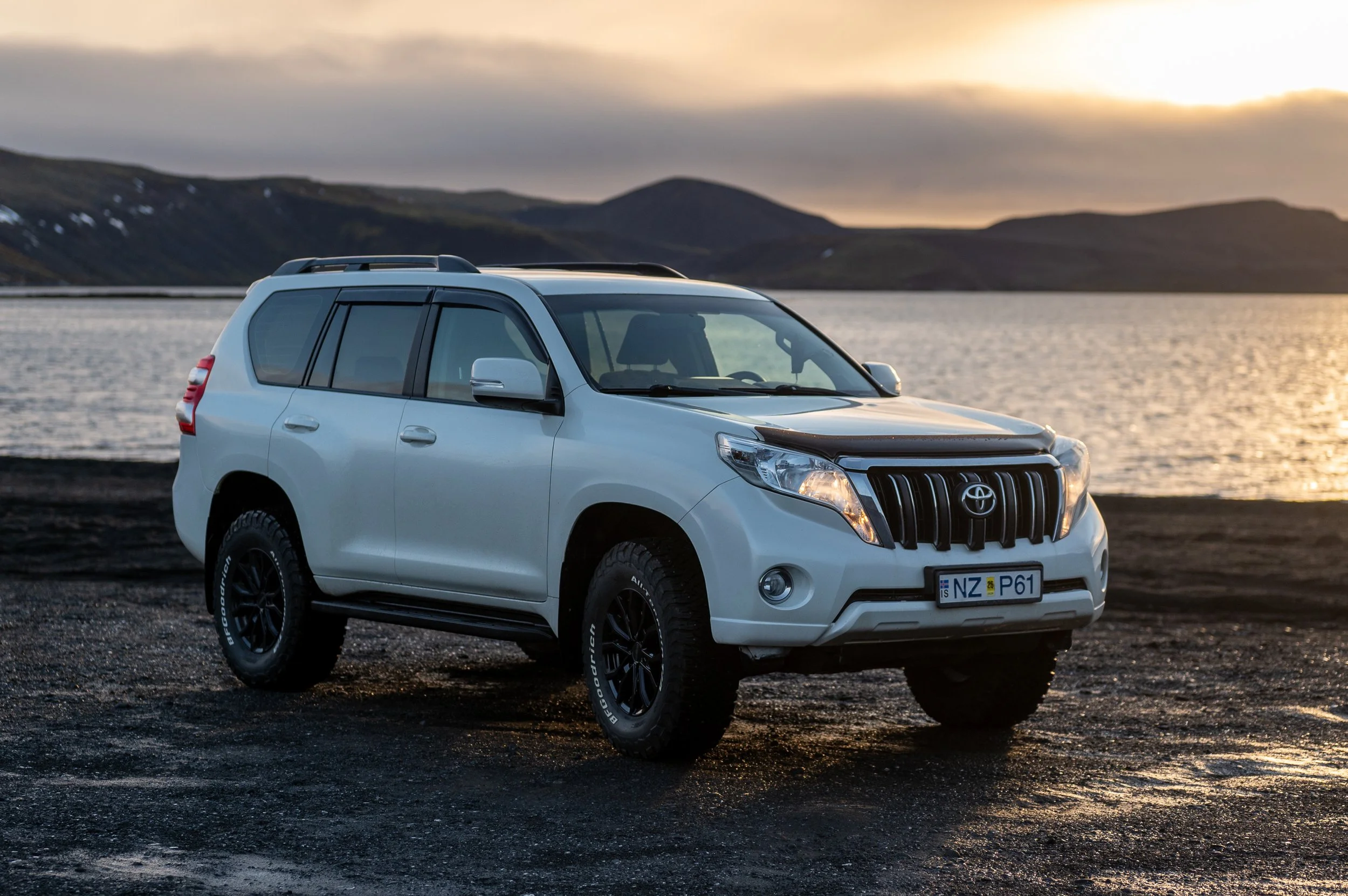 White SUV parked on a black gravel surface near a large body of water with mountains in the background during sunset.