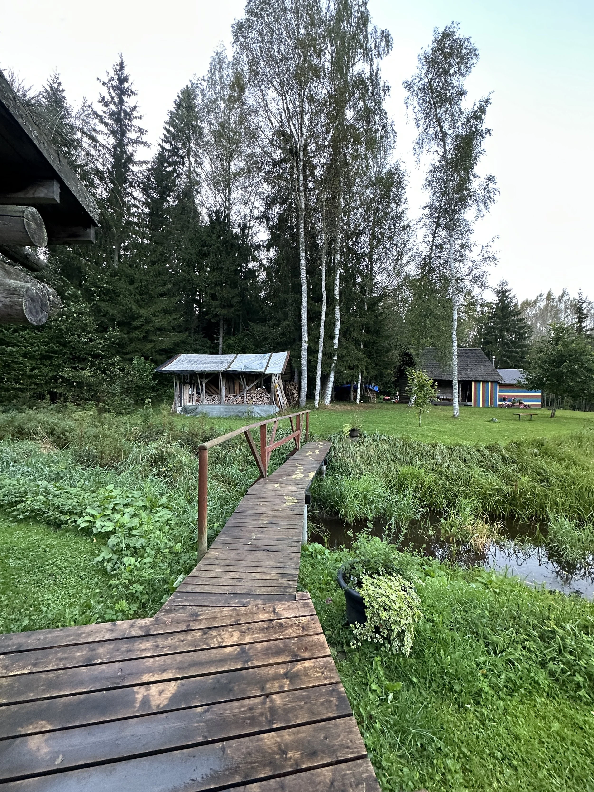 A rustic wooden footbridge crossing over a small pond or stream in a lush green yard, with trees and small buildings in the background.