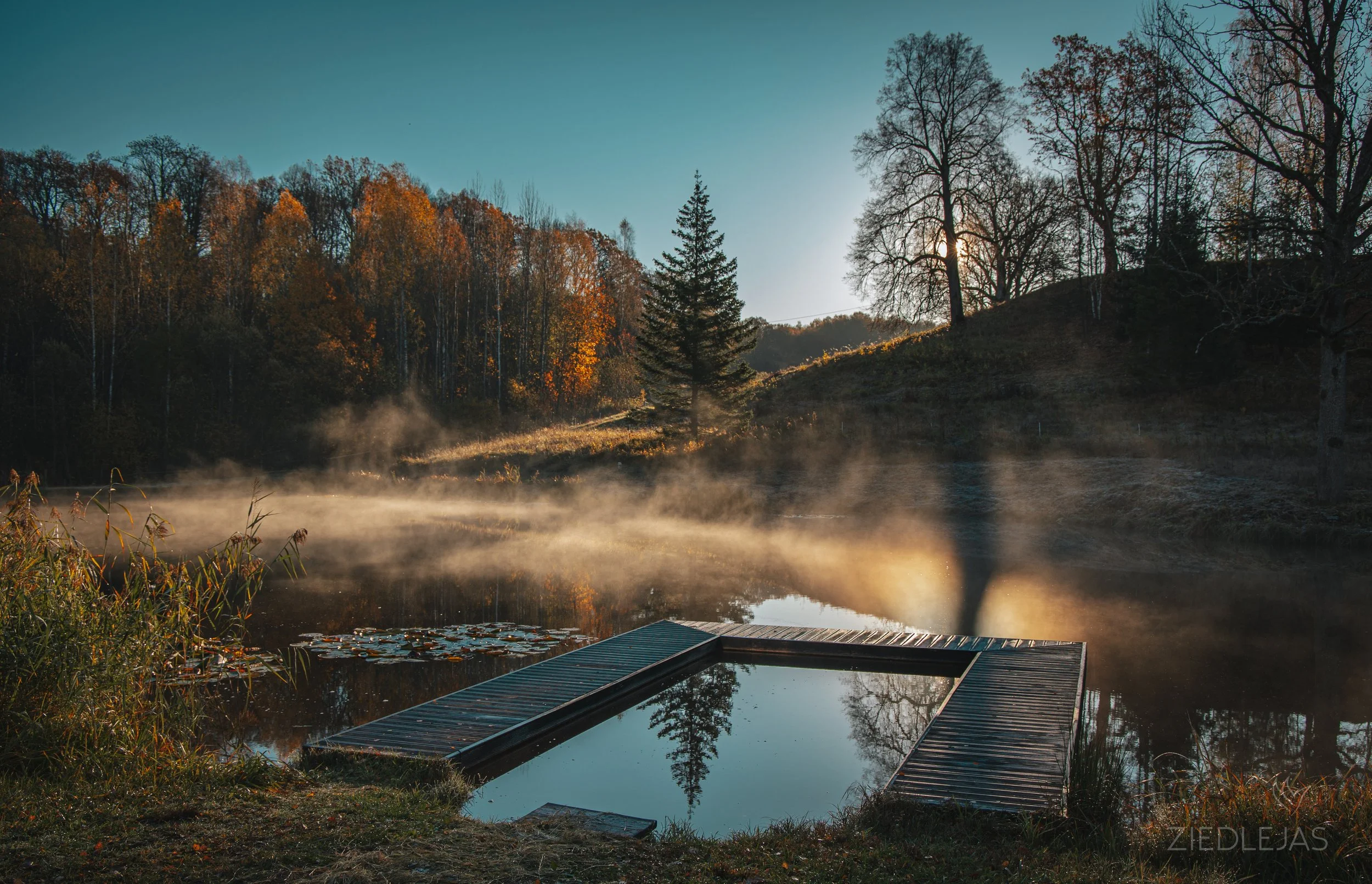 A calm lake at sunrise with mist rising from the water, surrounded by trees with fall foliage; a small dock extends into the lake, and the reflection of trees and the sky is visible in the water.