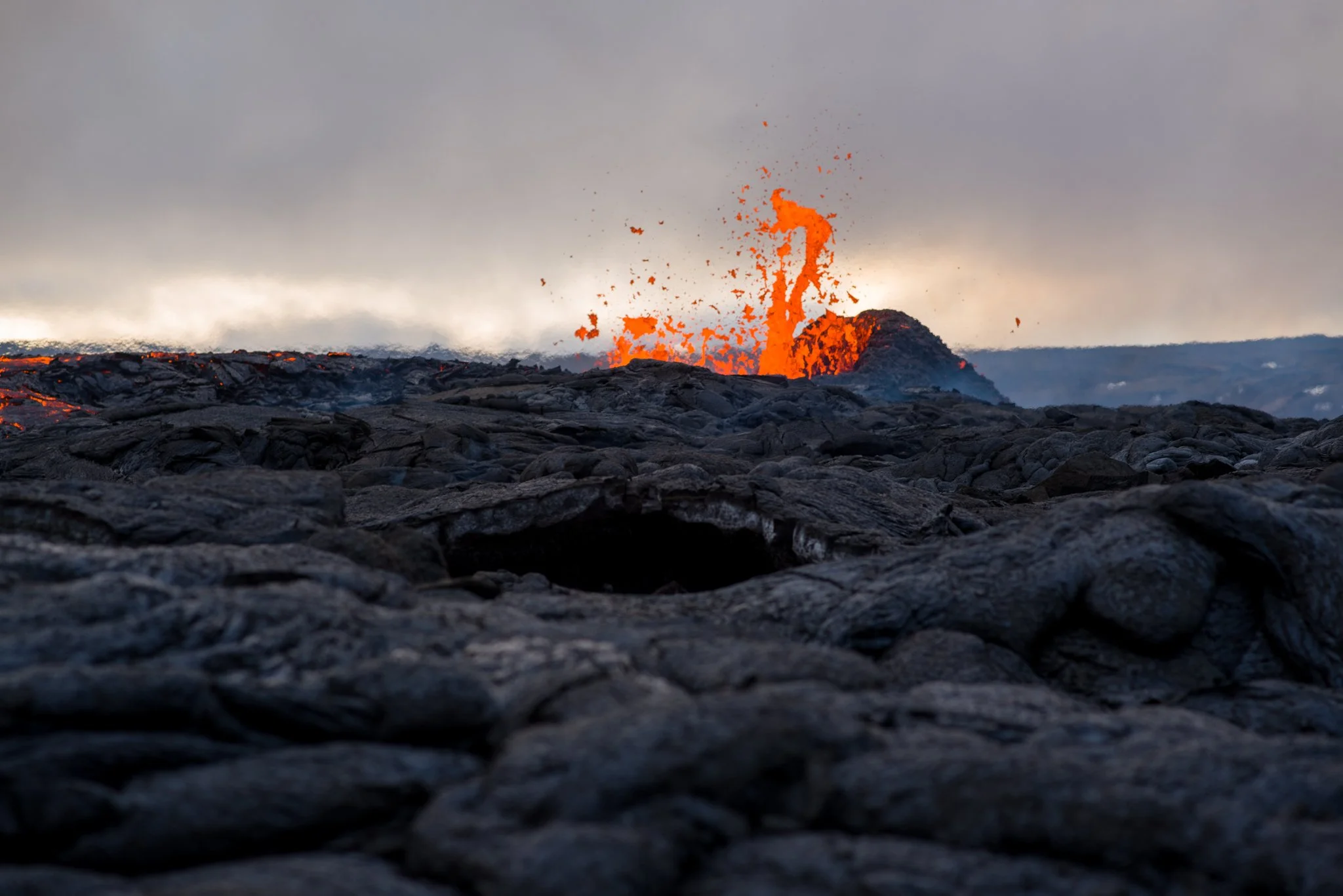 Eruption of lava spewing from an volcano on a dark, rocky landscape with a cloudy sky in the background.