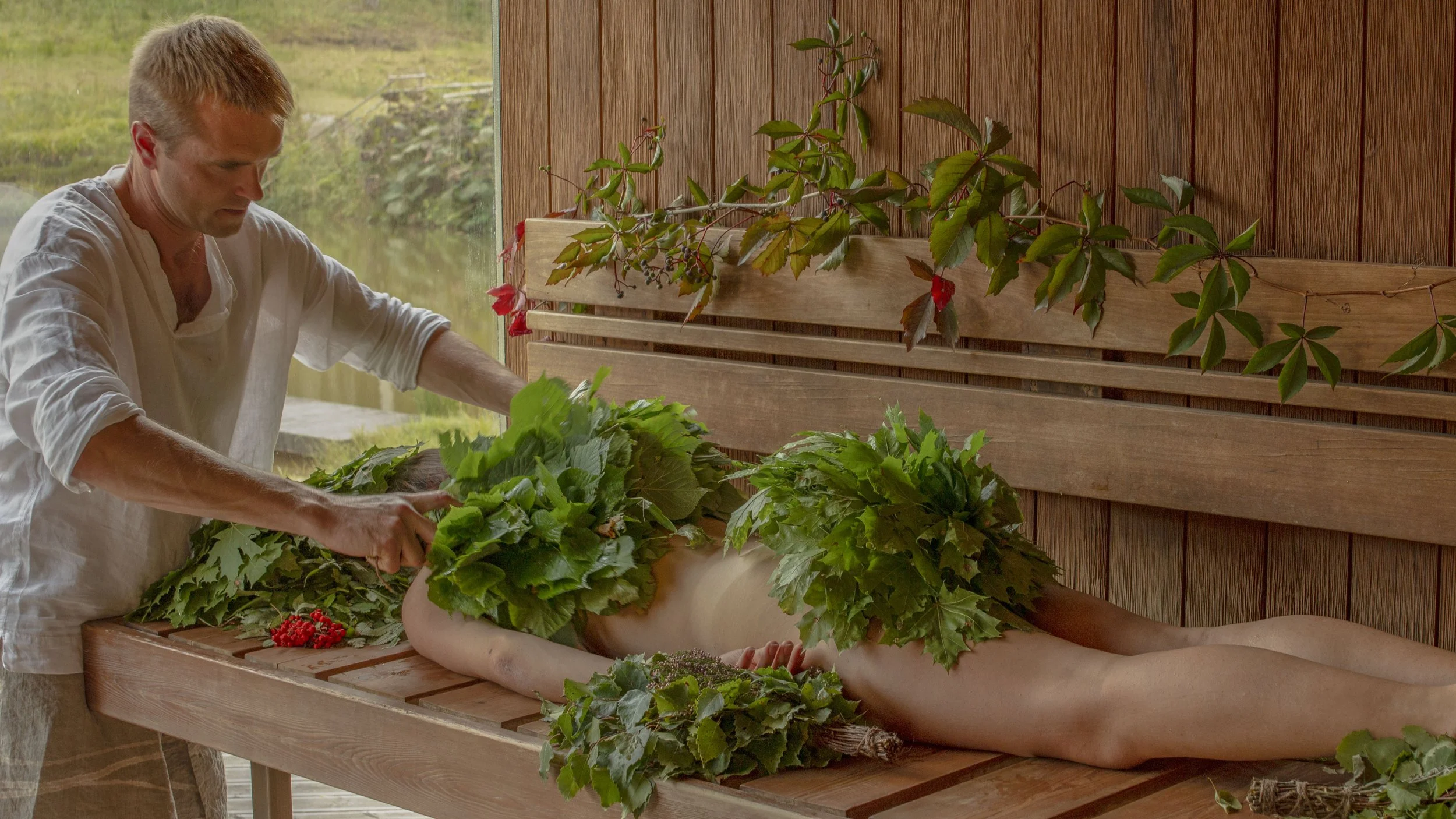 A person lying on a wooden table with leaves covering their body, while another person is tending to them in a room with wooden walls and a window.