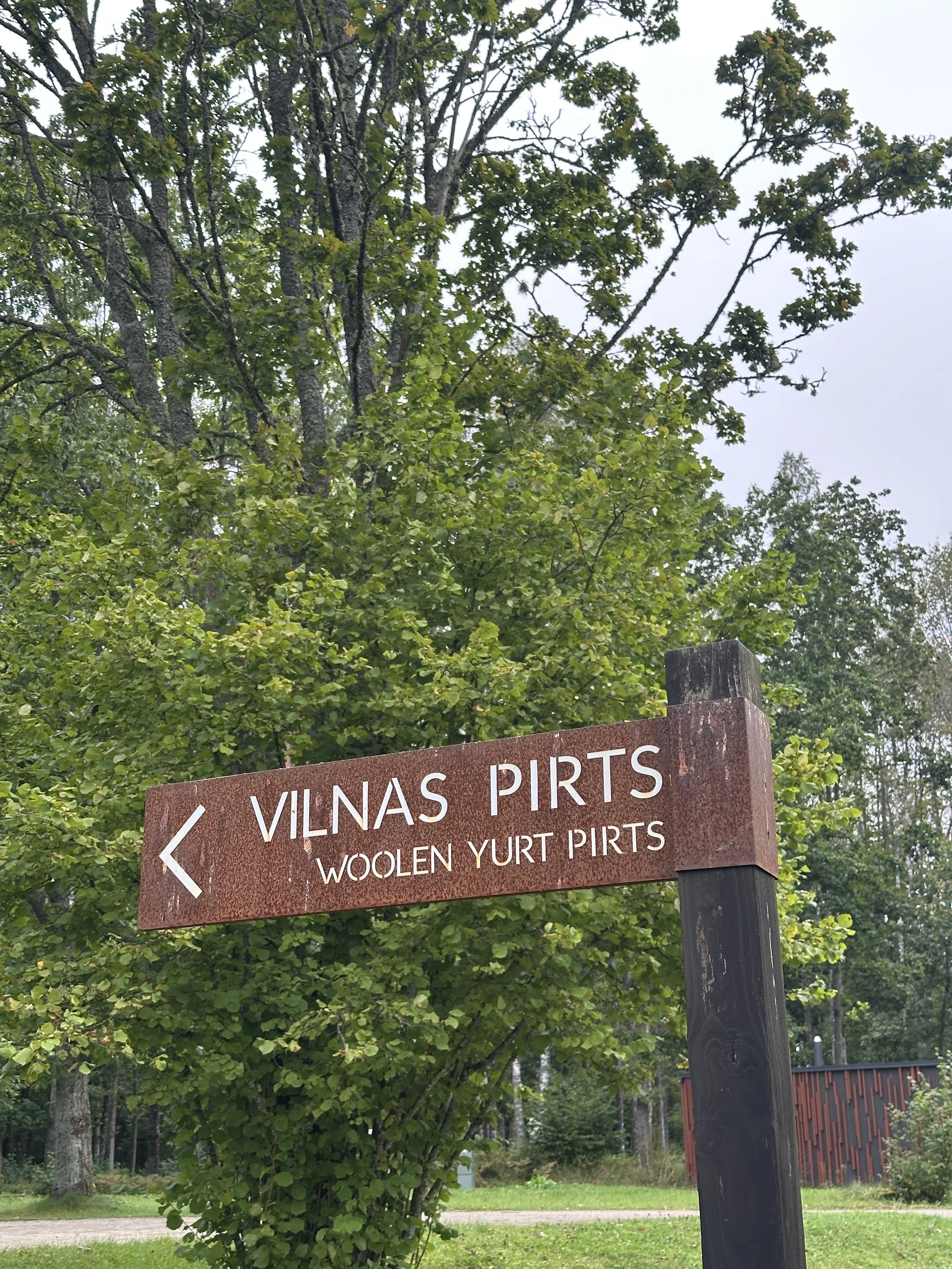 A wooden directional sign pointing left for Vilnas Pirts, which offers woolen Yurt Pirts. The sign is mounted on a dark wooden post with green trees and a building in the background.