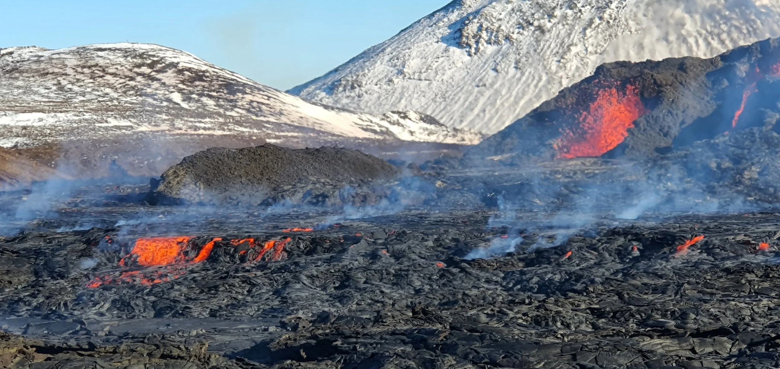 Erupting volcano with flowing lava, dark volcanic rocks, and snow-covered mountains in the background.