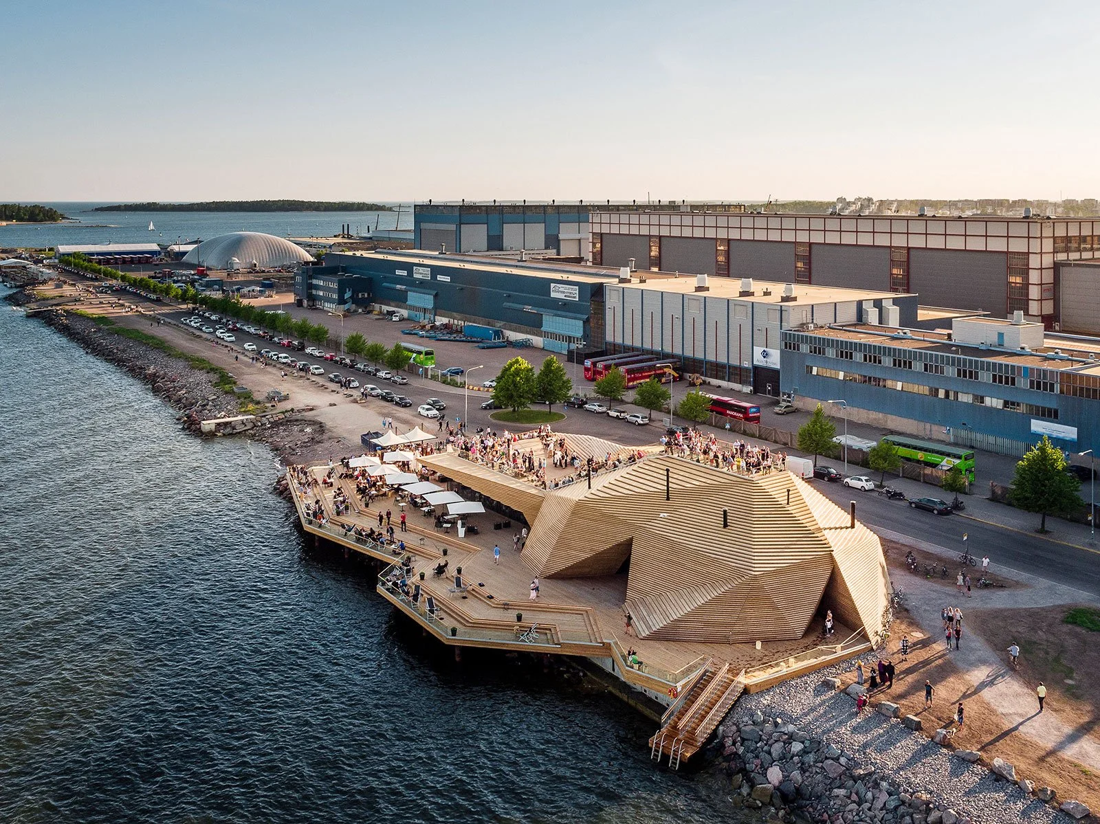 Aerial view of a waterfront park with a unique wooden pavilion, people walking and relaxing, with a parking lot and large industrial warehouse buildings nearby.
