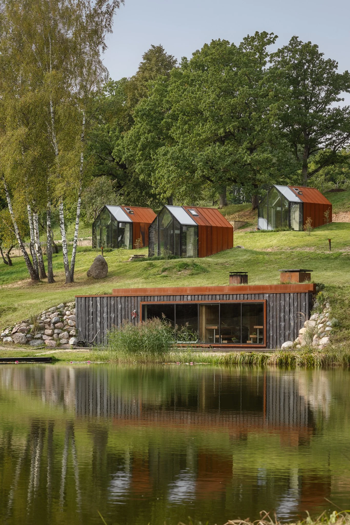 Eco-friendly glass and wood tiny houses on a lush green hillside near a pond, surrounded by trees and rocks.