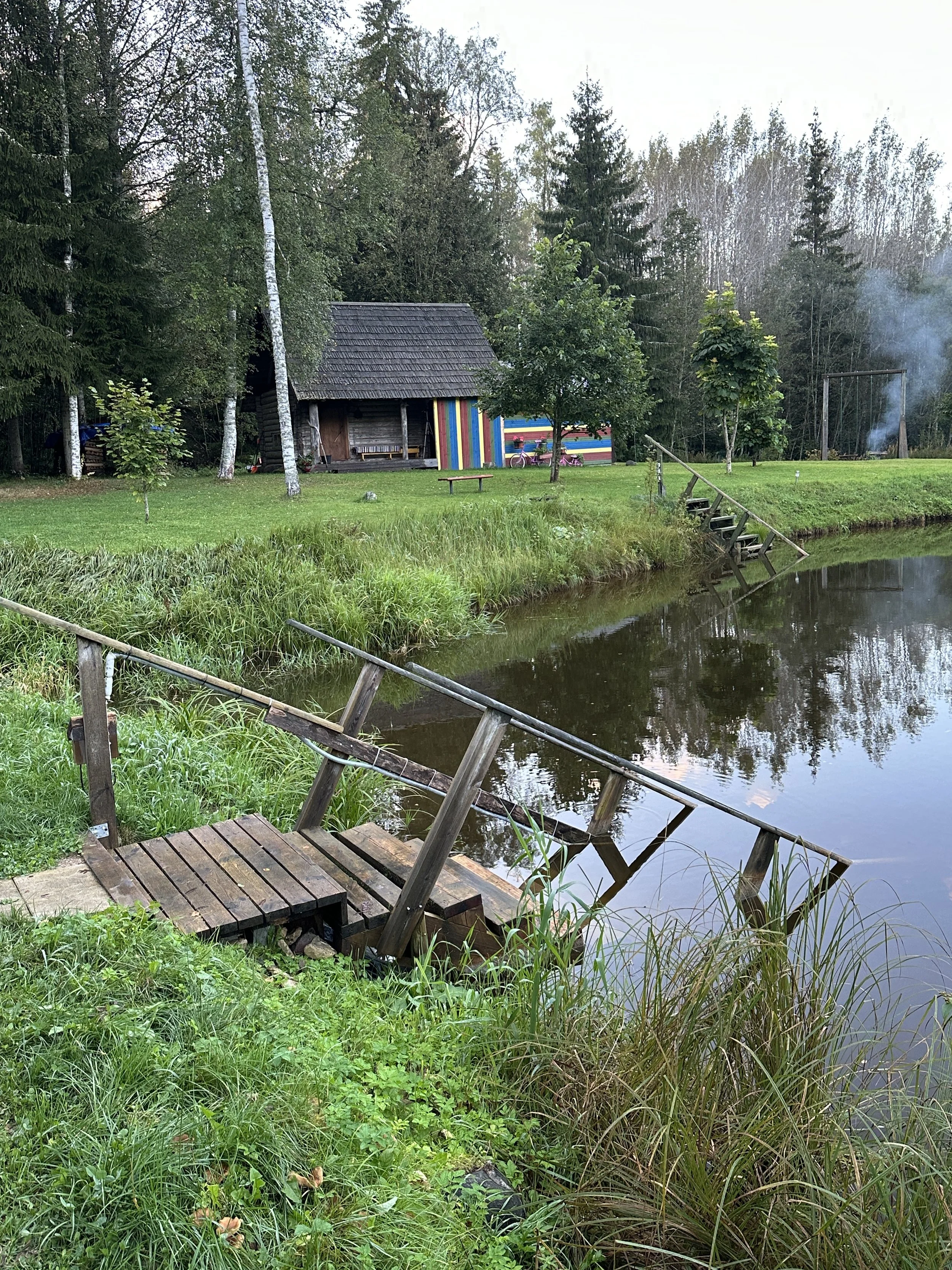 A lakeside scene with a small, tilted wooden dock in the foreground, grassy shoreline, a calm body of water, a rustic wooden house with a collapsible front porch, and colorful striped panels in the background. There are trees, a bench, and a swing se