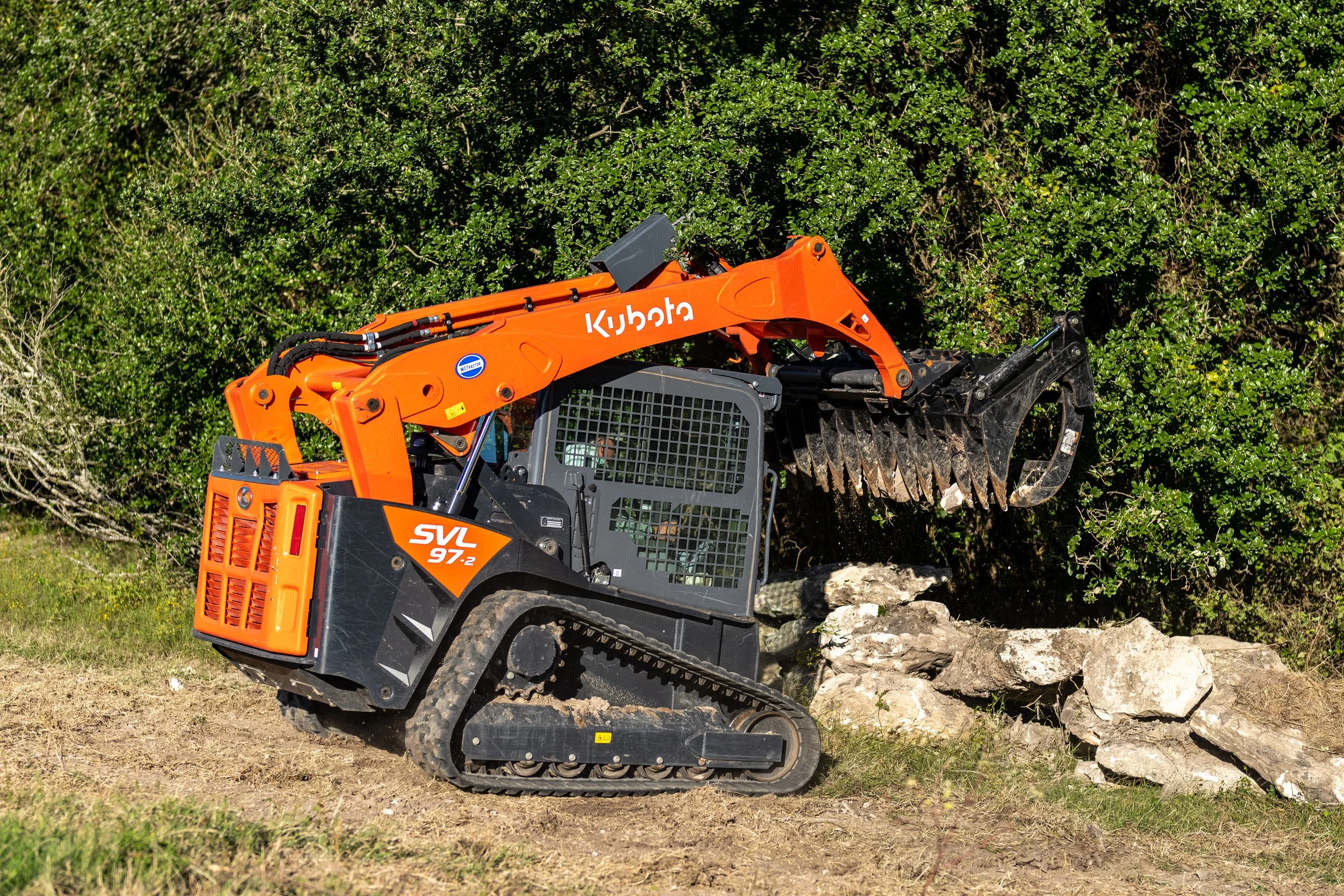 Kubota SVL97-2 compact track loader moving rocks in a grassy area with trees in the background.