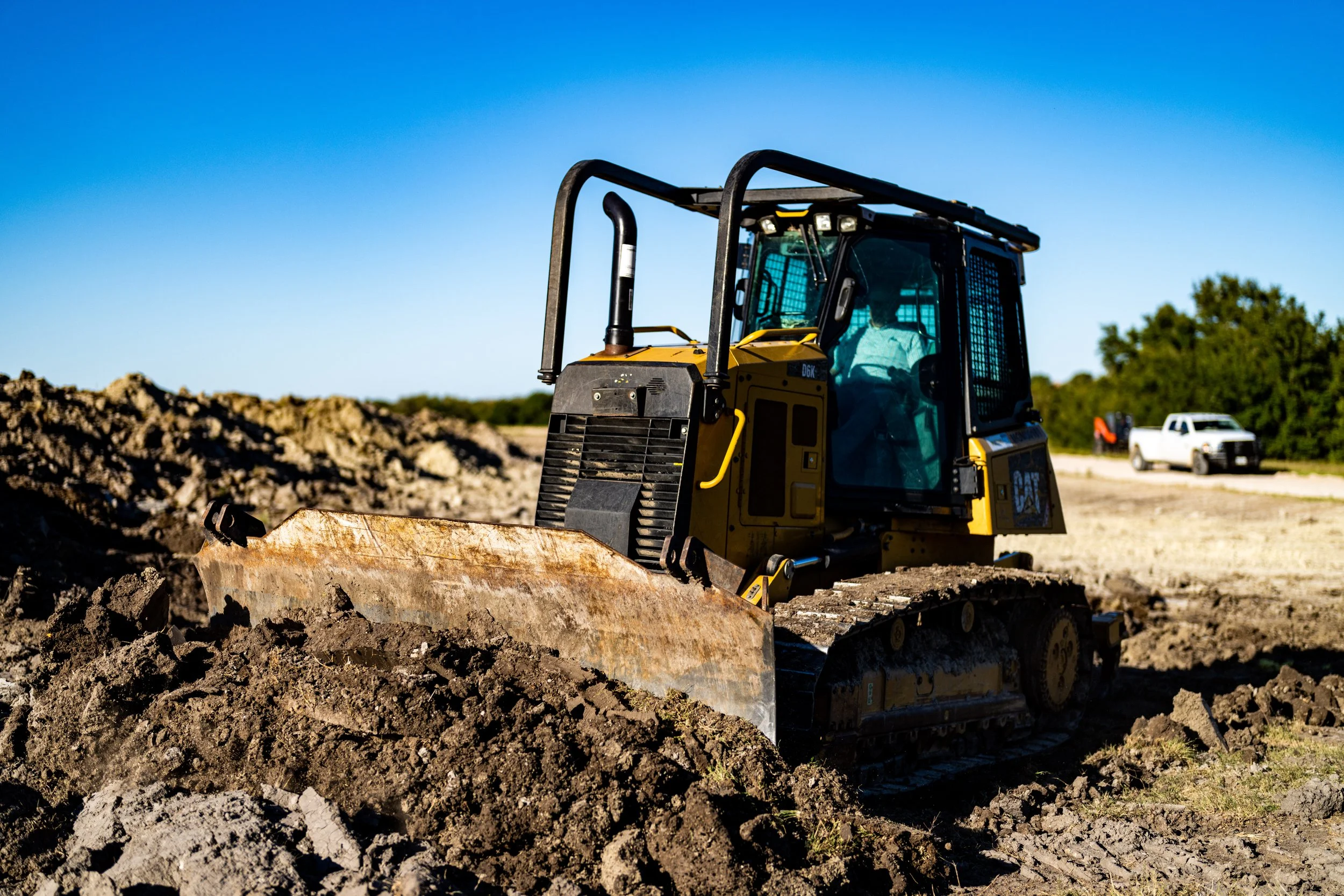 A yellow bulldozer moving earth at a construction site, with a blue sky background and a white truck in the distance.