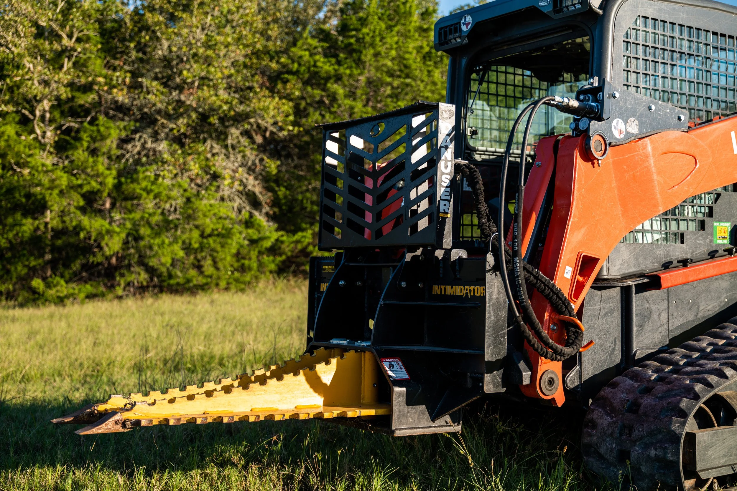 Skid steer loader equipped with a tree puller attachment on grassy terrain in a wooded area.