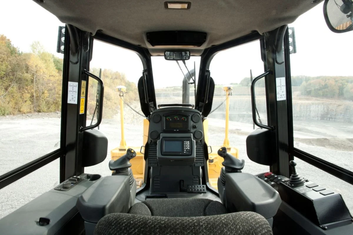 Interior view of a heavy construction vehicle, showing the control panel and joystick controls with a view of an excavation site outside.