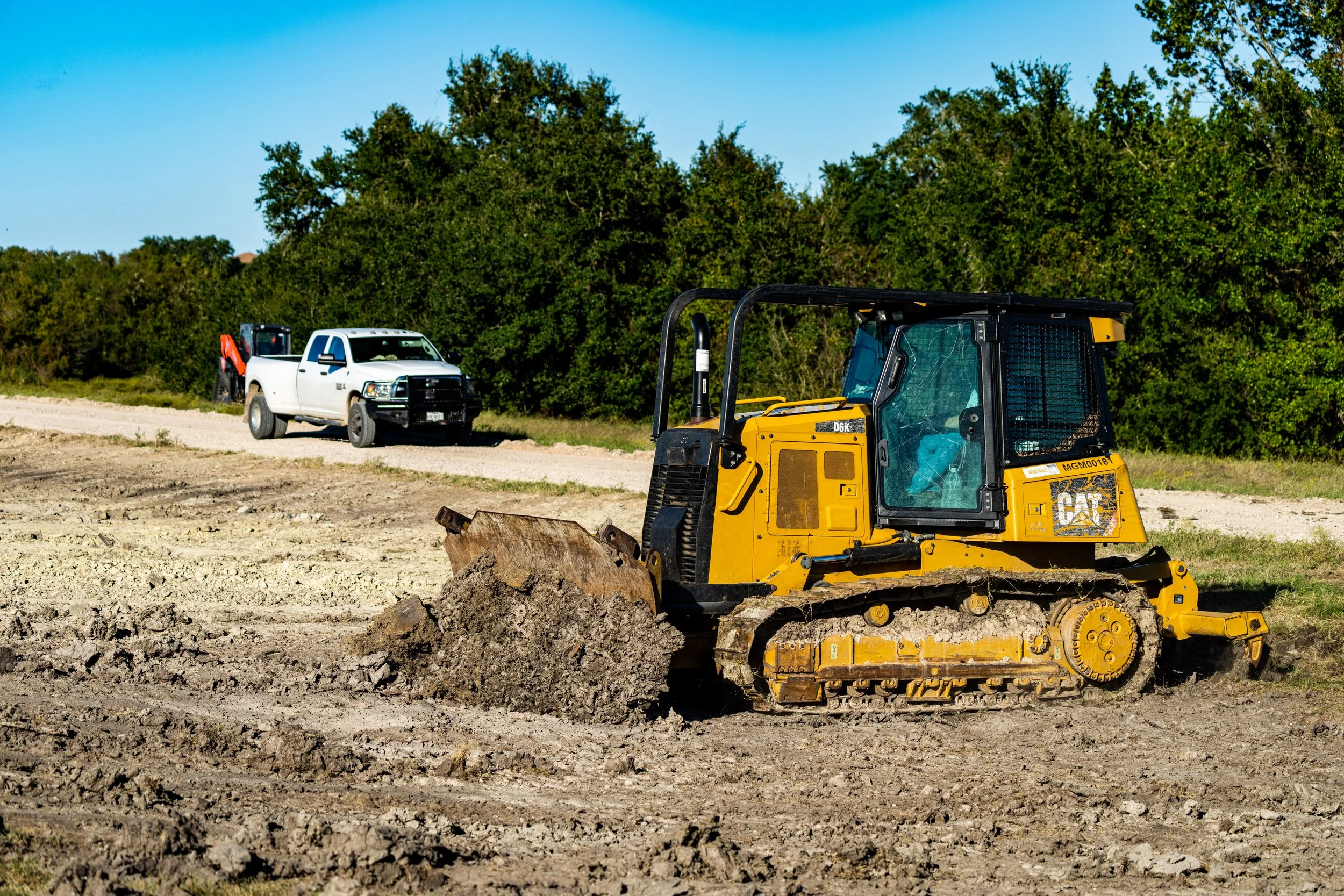 A yellow bulldozer pushing dirt on a construction site, with a white pickup truck in the background, surrounded by trees under a clear blue sky.