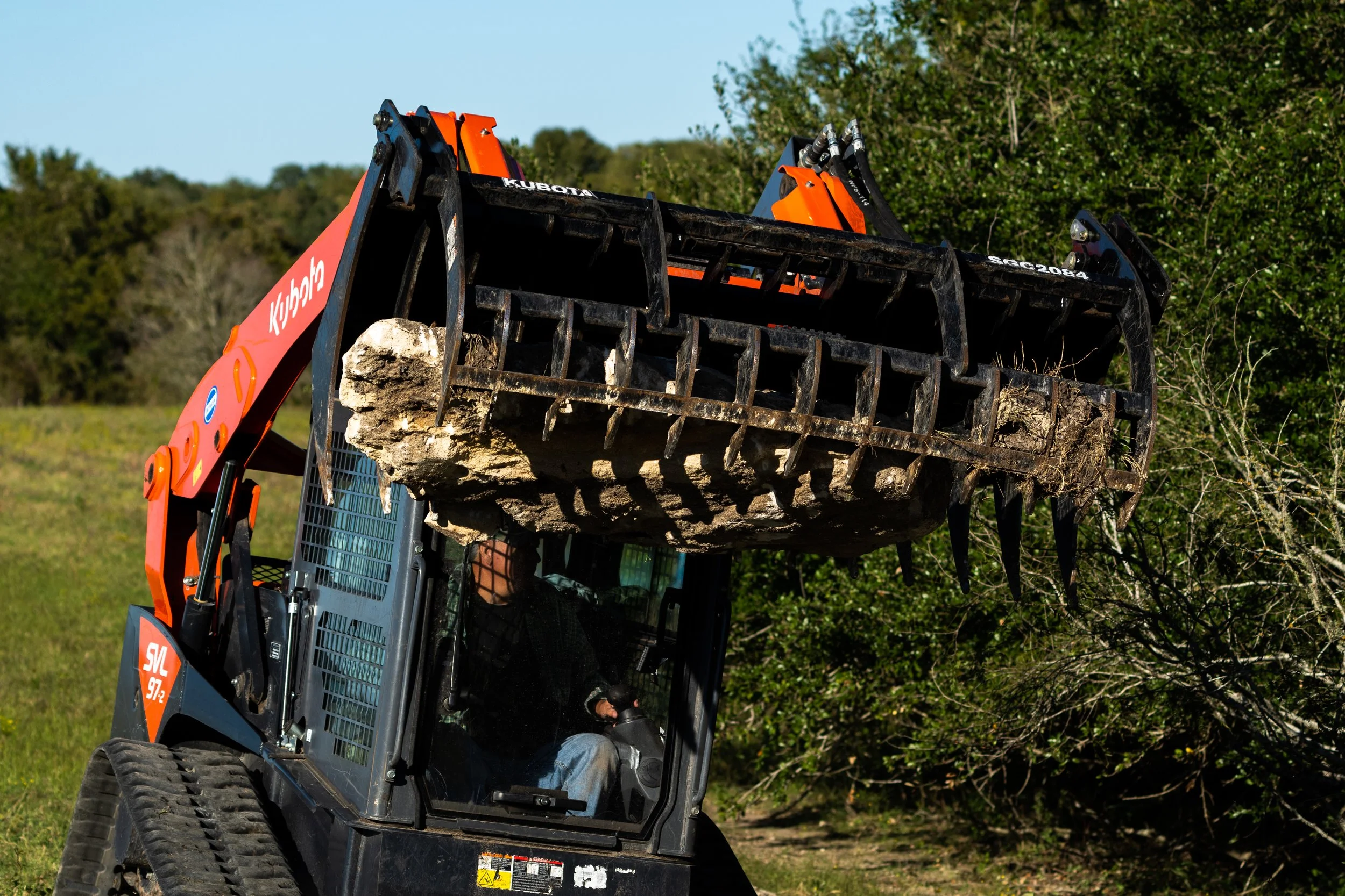 A Kubota skid steer loader lifting a large rock with a grapple attachment in a grassy area.
