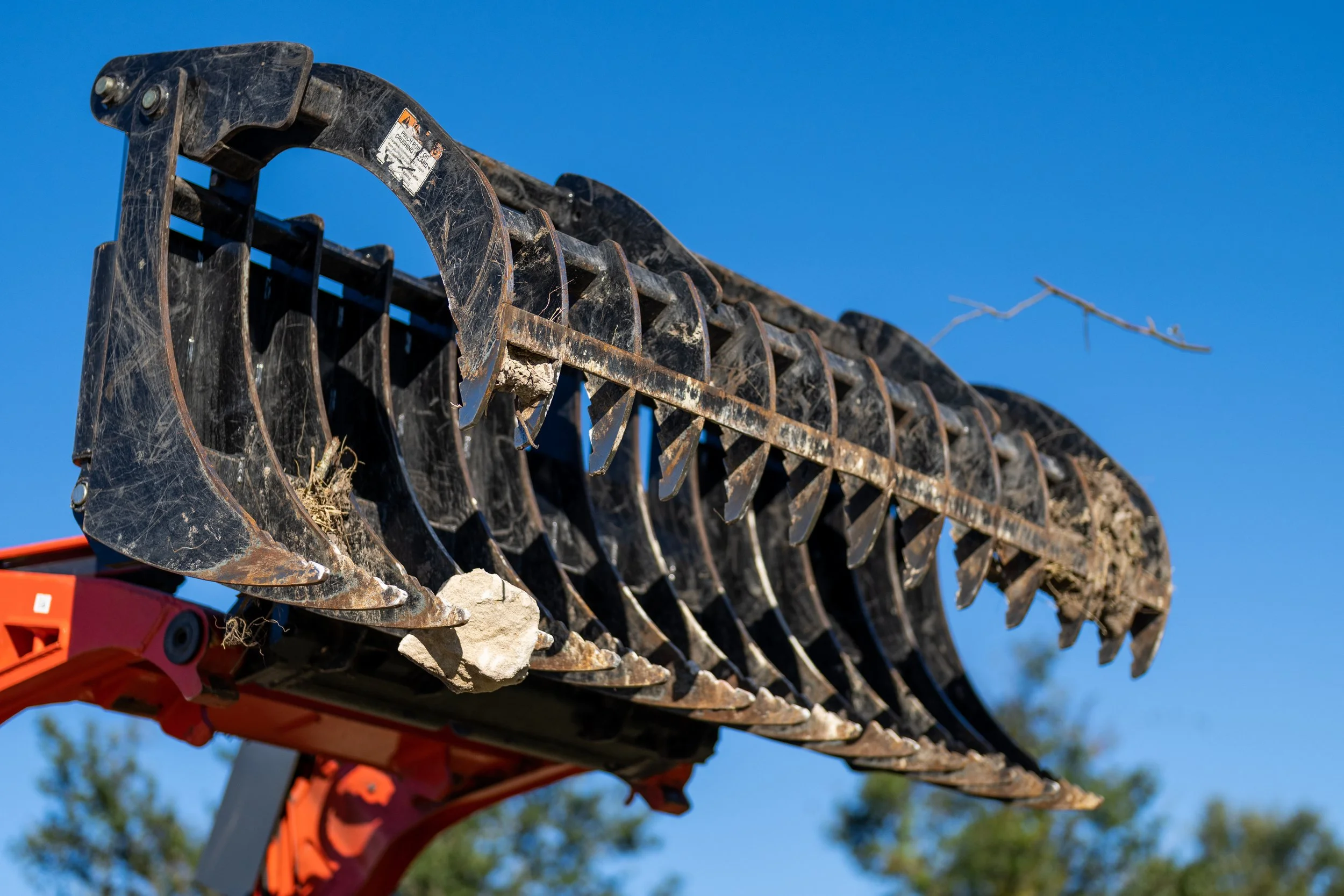 Close-up of a large construction machinery grapple claw with dirt and rocks, against a clear blue sky.