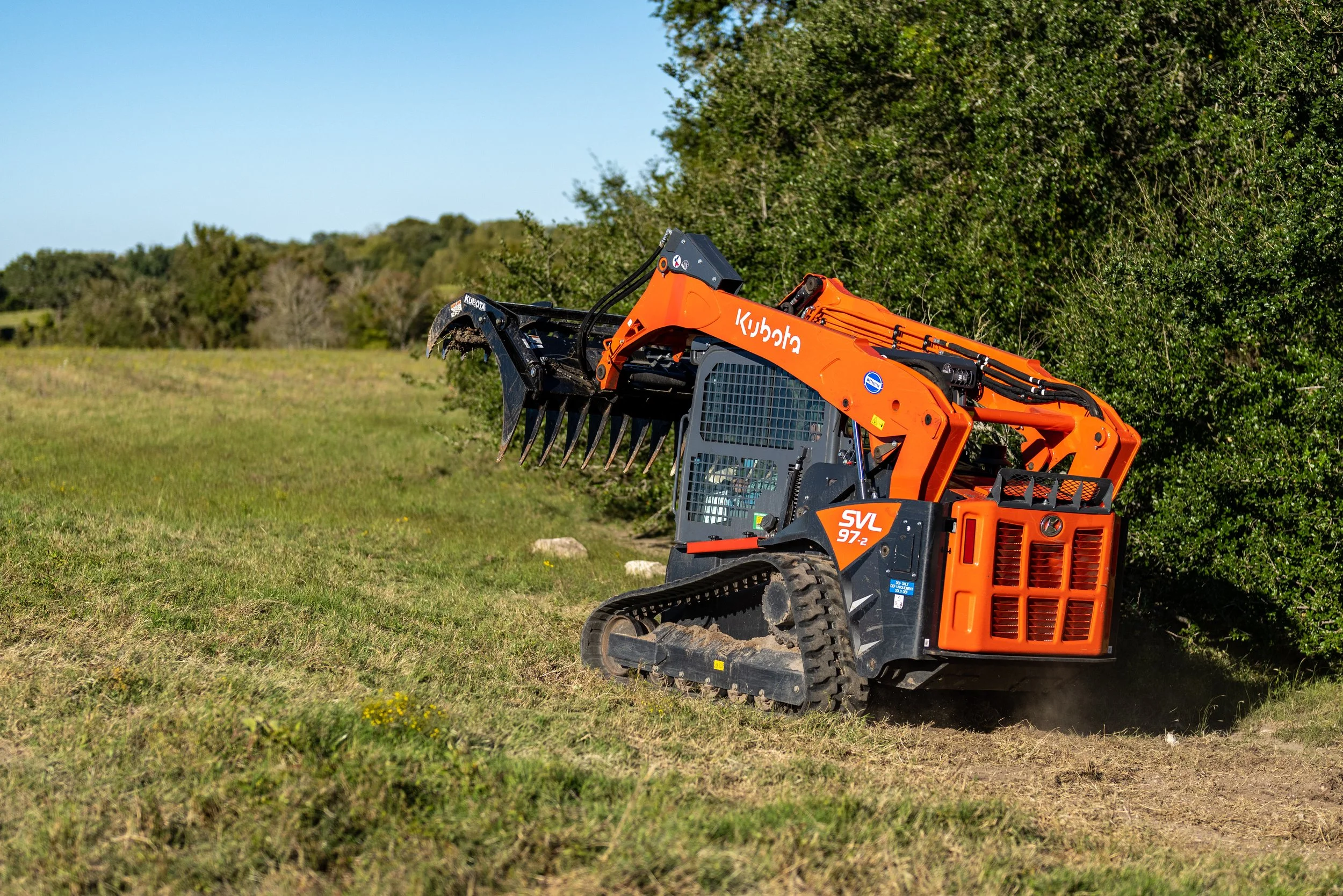 Orange Kubota SVL97-2 compact track loader with a grappler attachment on grassy terrain, surrounded by trees, under a clear blue sky.